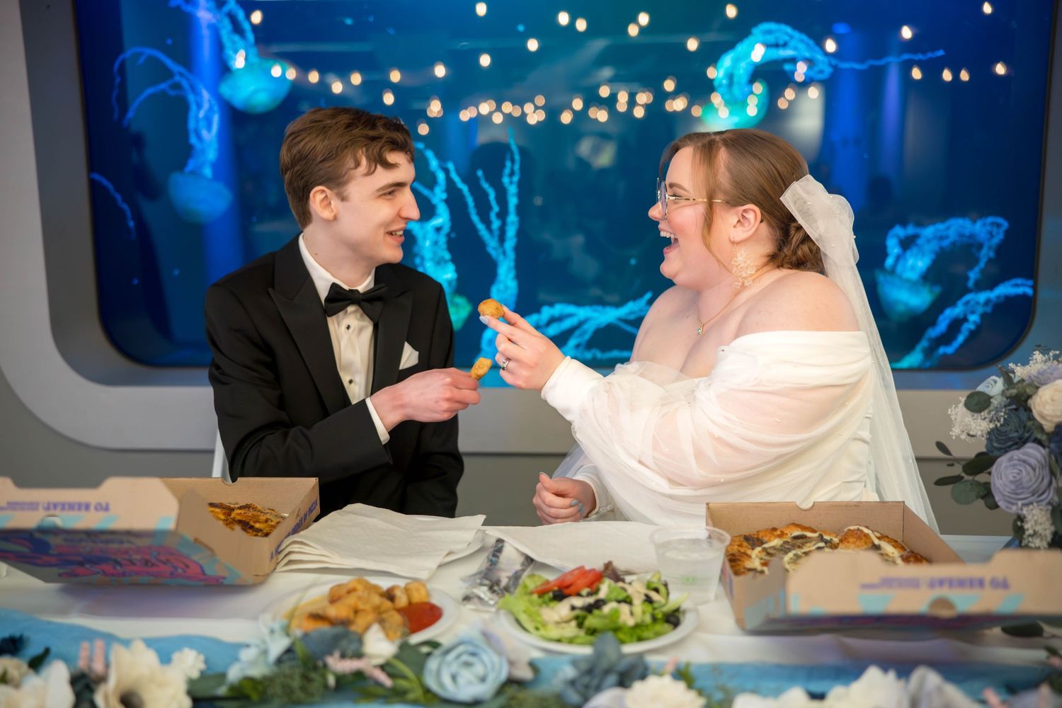 Newlyweds share a playful moment while dining at a reception with string lights and food platters.