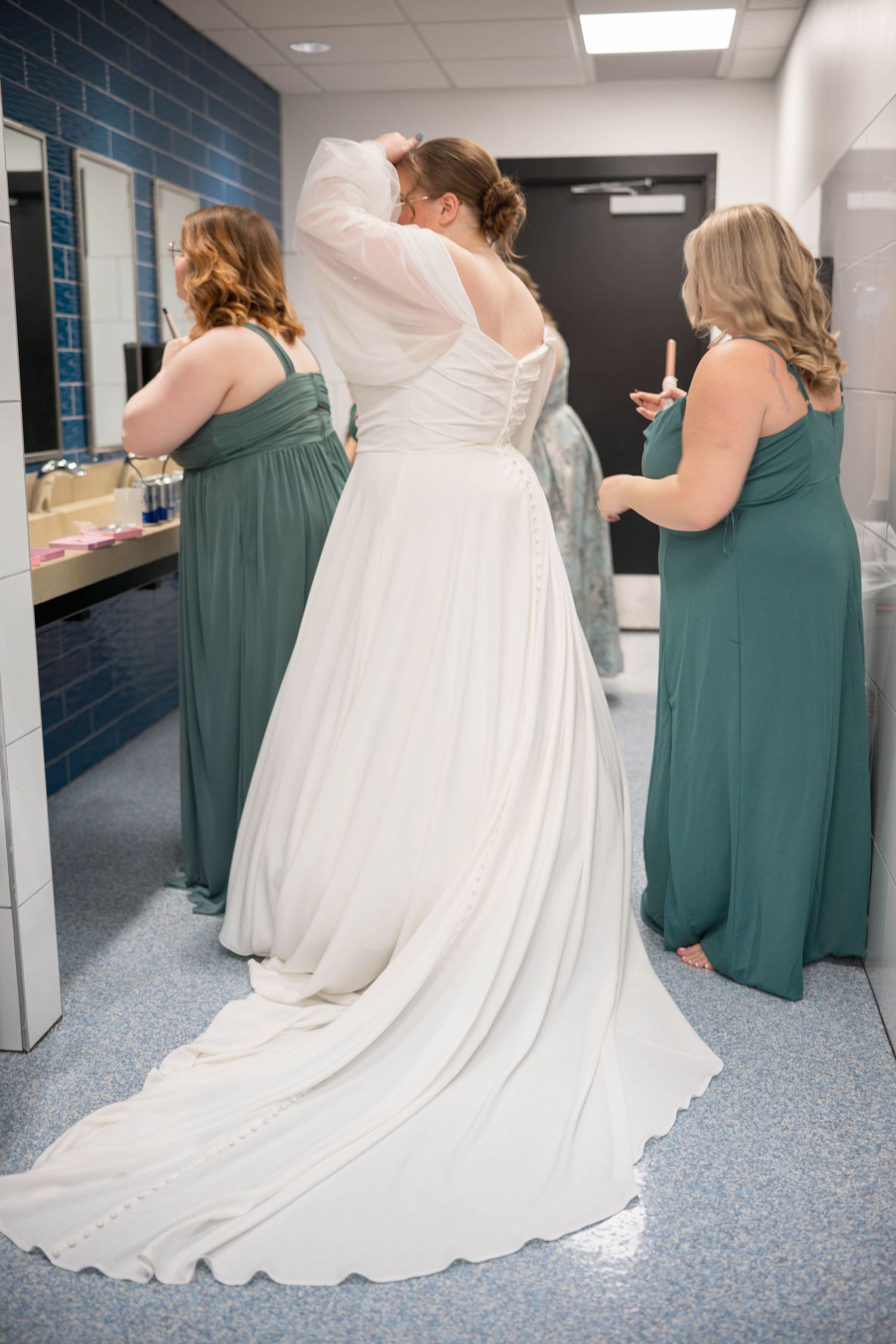 A bride in a flowing white wedding gown gets assisted by bridesmaids wearing sage green dresses in a preparation room.