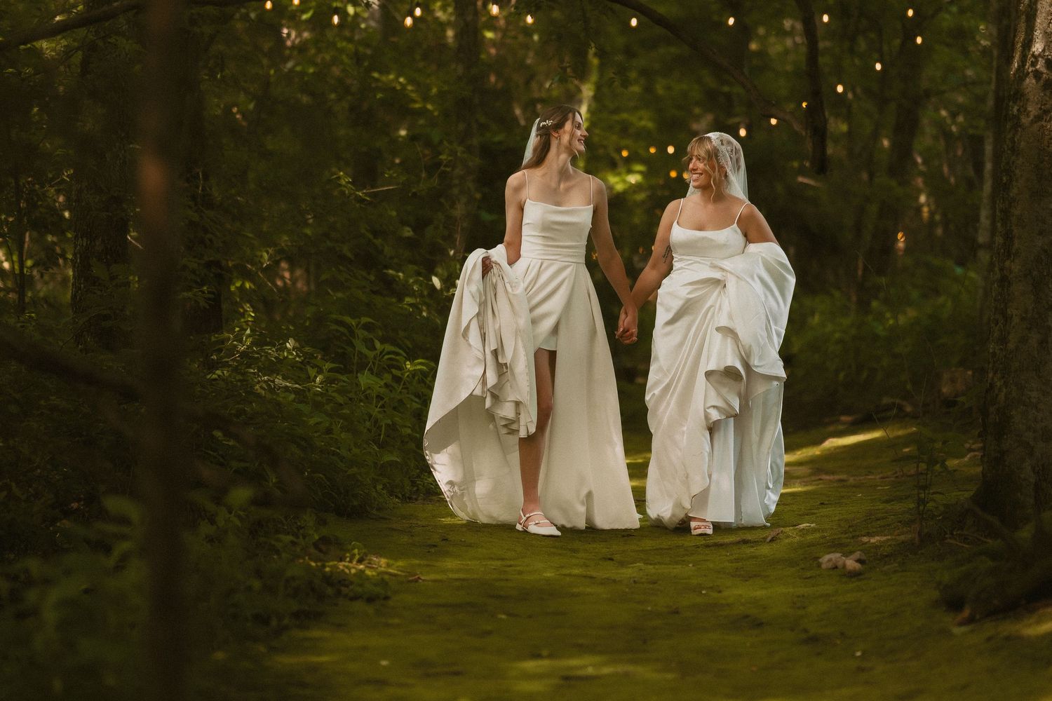 Two people in flowing white wedding dresses walk together along a sunlit forest path during a magical outdoor ceremony.