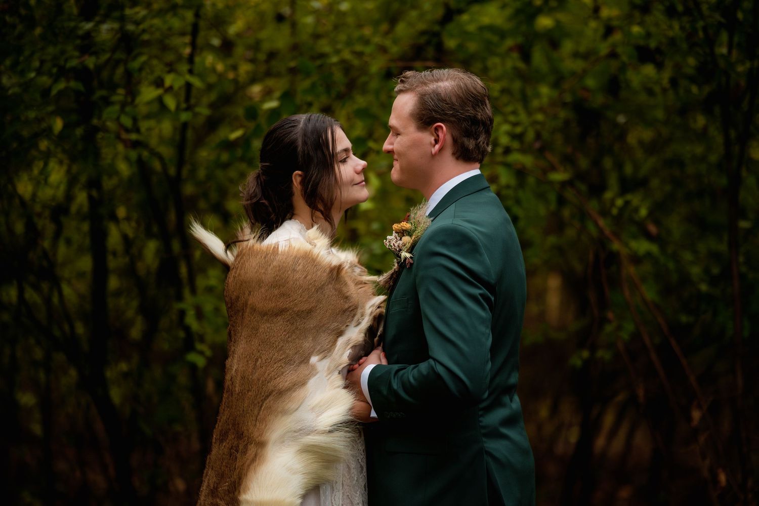 A couple in wedding attire embraces in a wooded outdoor setting at sunset.