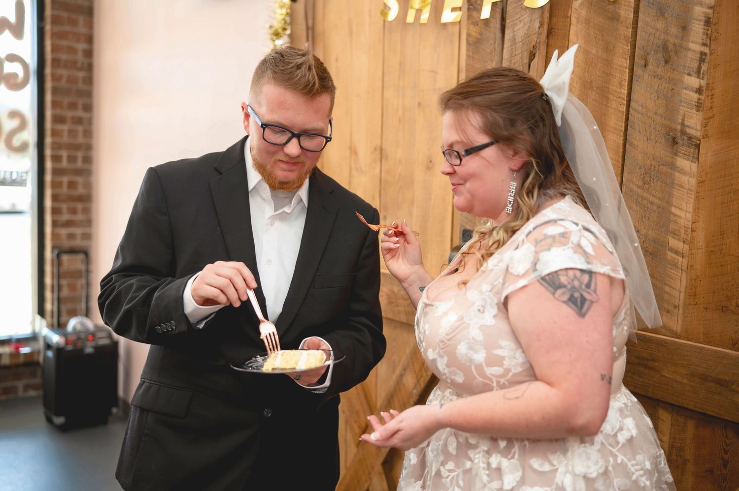 A couple shares cake during their wedding reception in a rustic venue with brick walls and wooden decor.