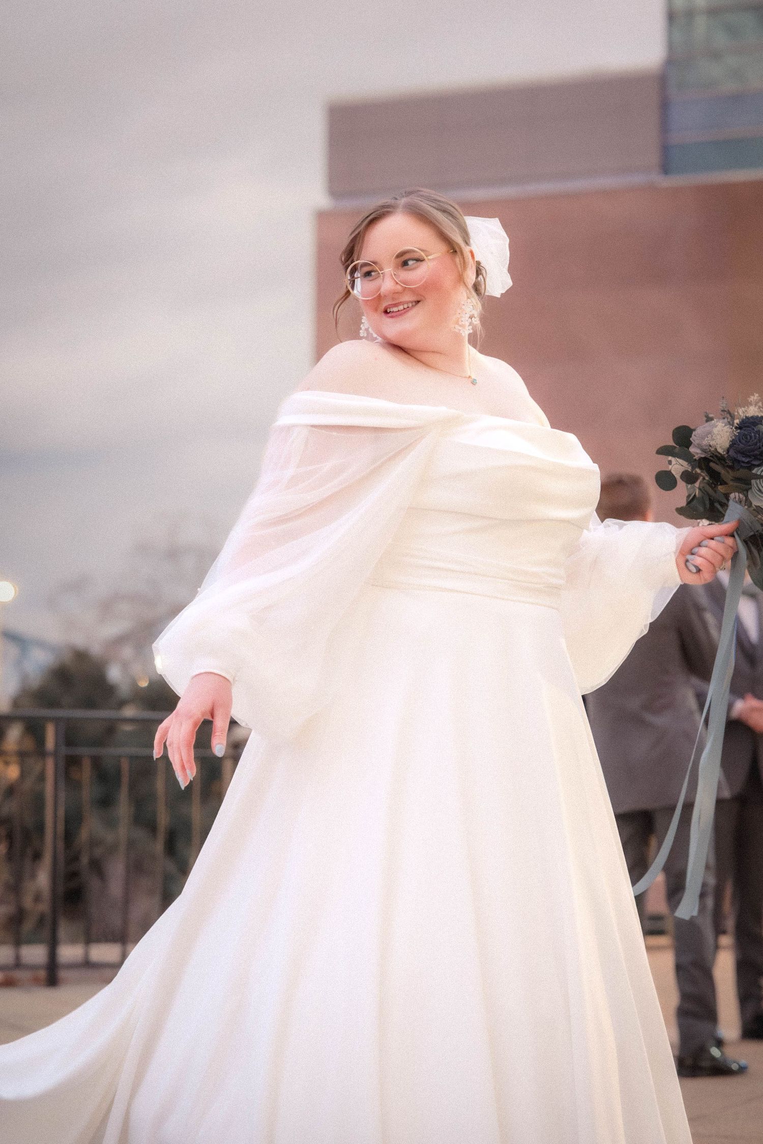 A bride in a flowing white wedding dress and shawl poses outdoors on a balcony with mountains in the background.