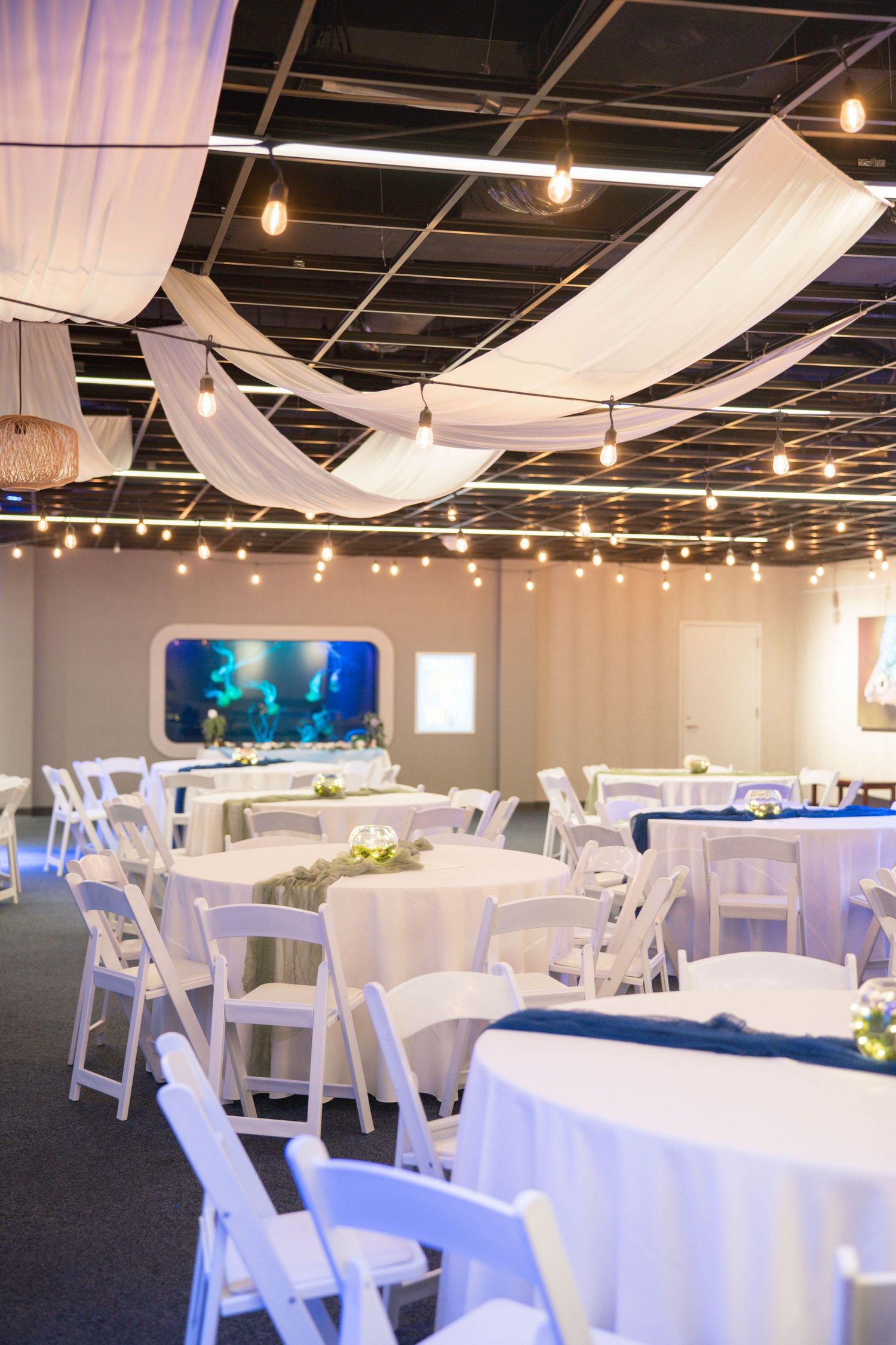 An elegant reception venue with white draped fabric ceiling, string lights, round tables, and white folding chairs.