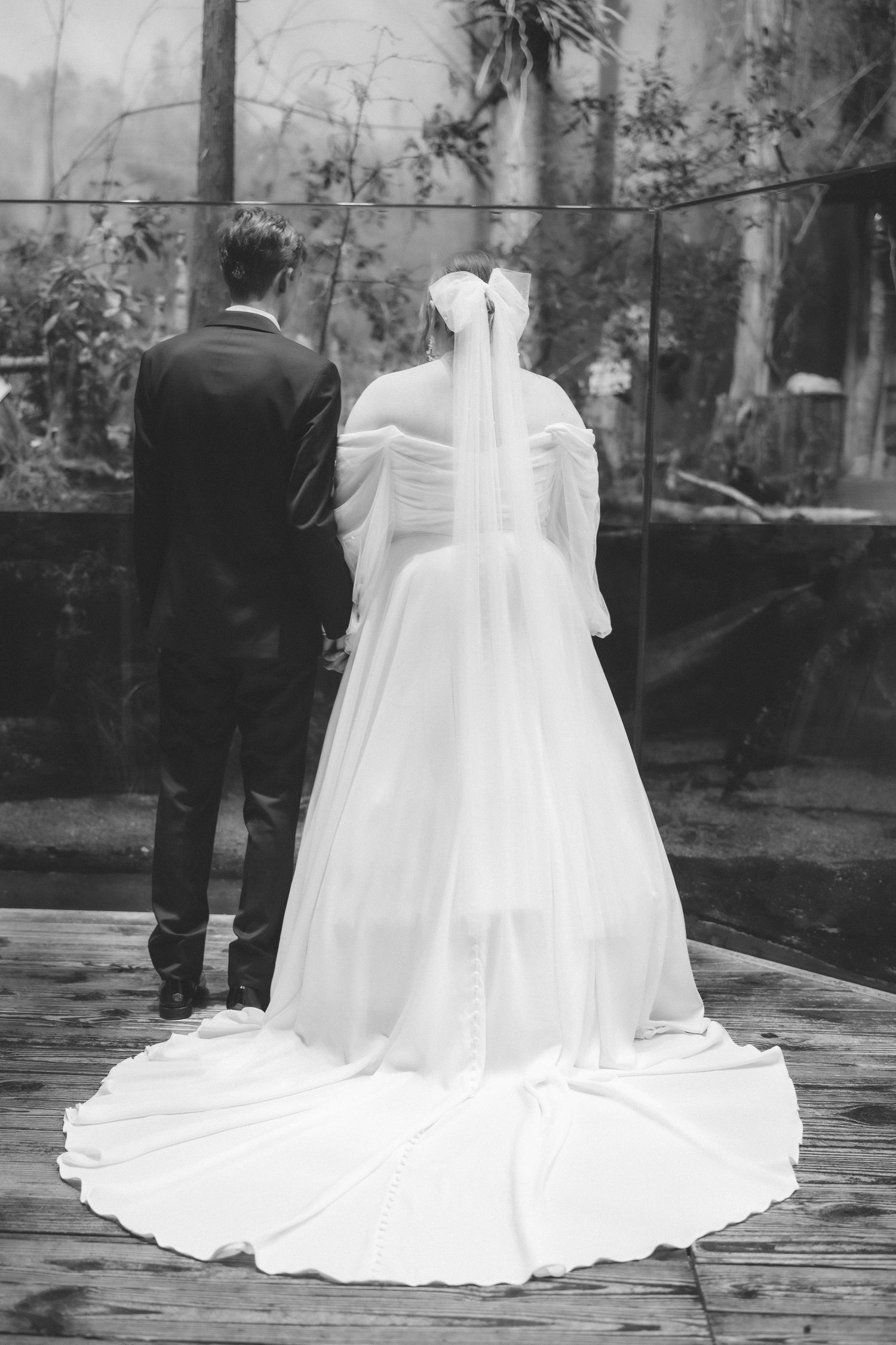 Black and white photo of a bride in a flowing white gown with long train standing next to a groom in formal attire.