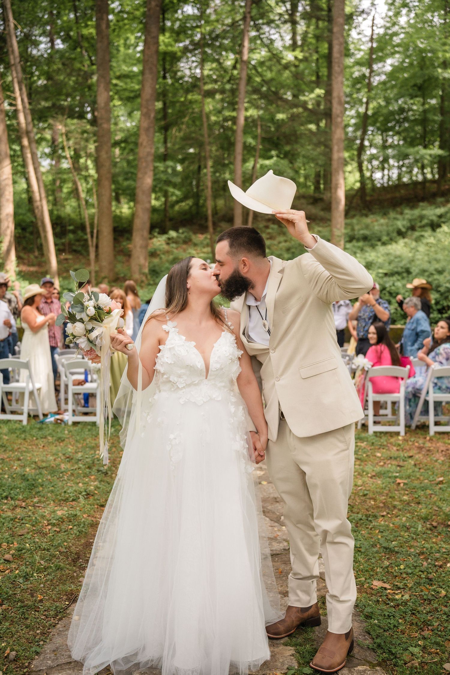 A couple in wedding attire kisses outdoors surrounded by wooden chairs and guests during a forest ceremony reception.