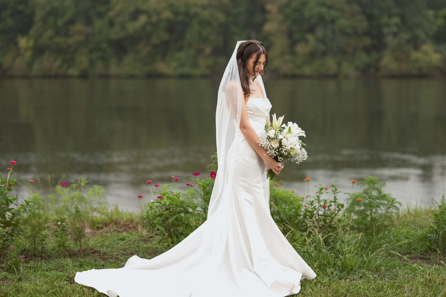 A bride in a flowing white mermaid dress and long veil holds a white bouquet while standing beside a serene lake.