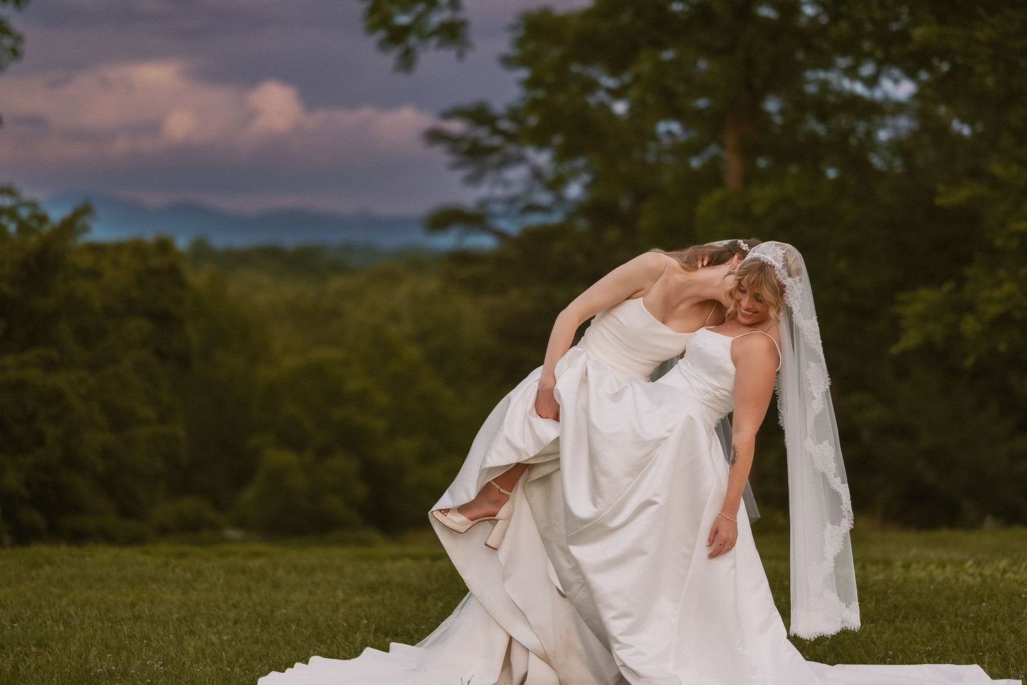 A bride in a flowing white wedding dress twirls outdoors at sunset against a scenic mountain backdrop.