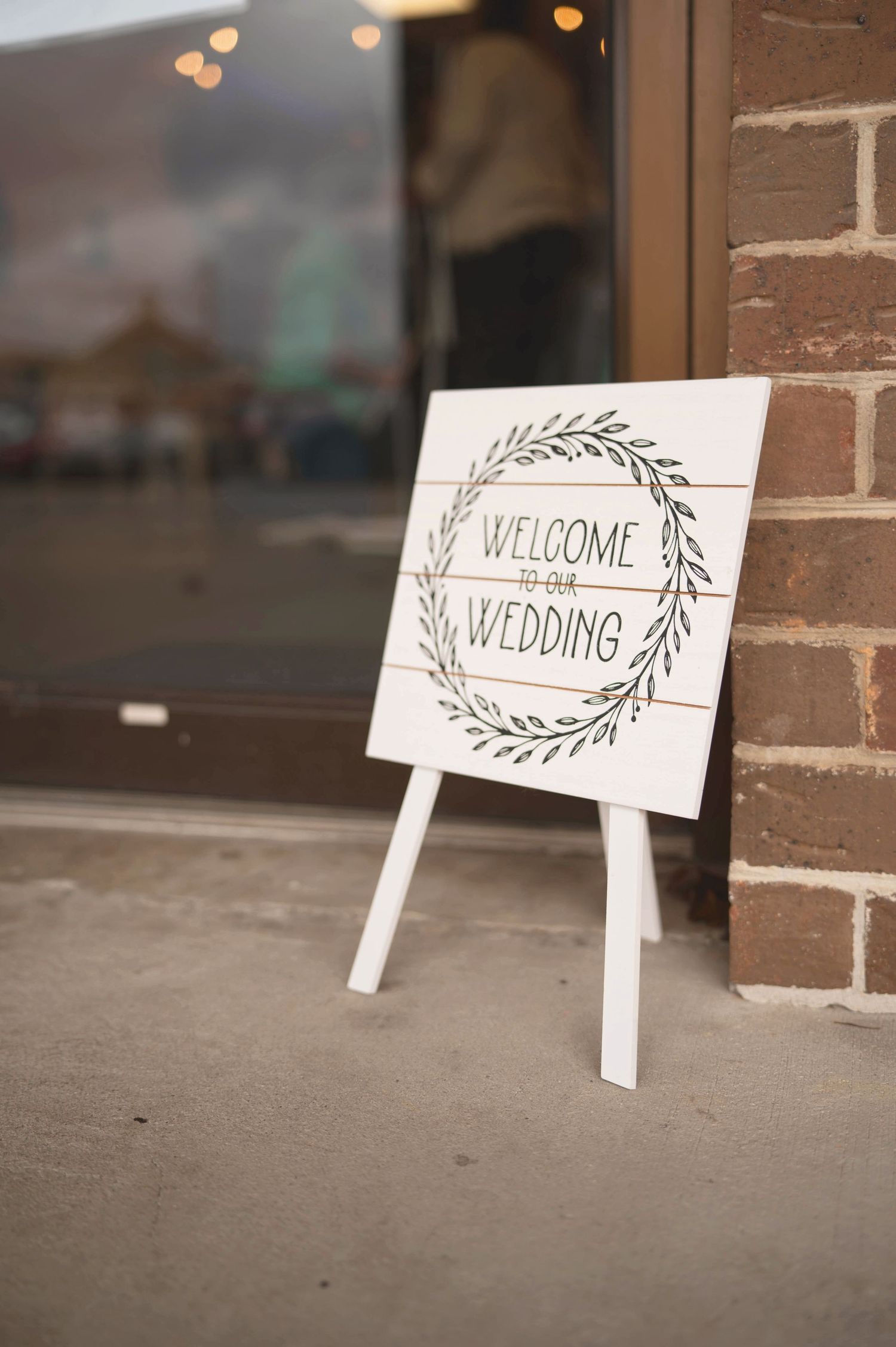 A white welcome wedding sign with a hand-drawn wreath design stands on a small easel against a brick wall.