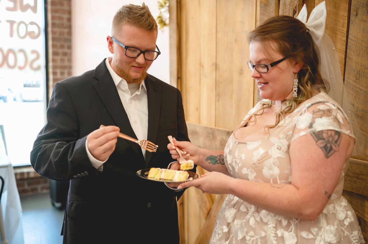 A couple in formal wedding attire share a slice of cake together at their reception against a wooden wall backdrop.