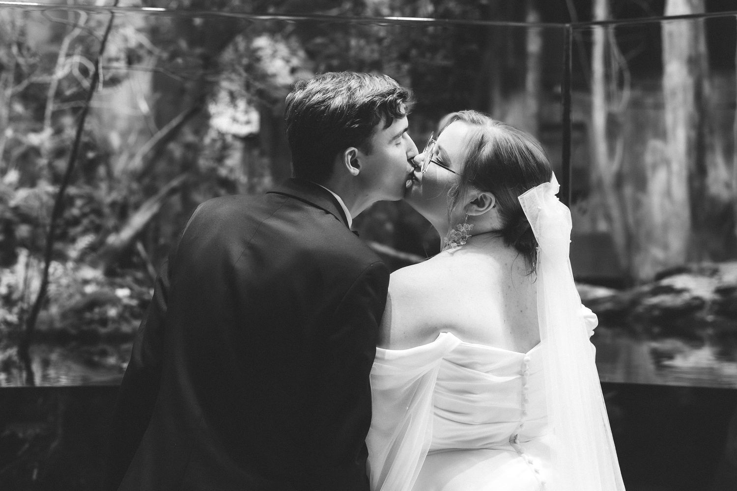 Black and white photo captures newlyweds kissing during intimate woodland wedding portrait session.