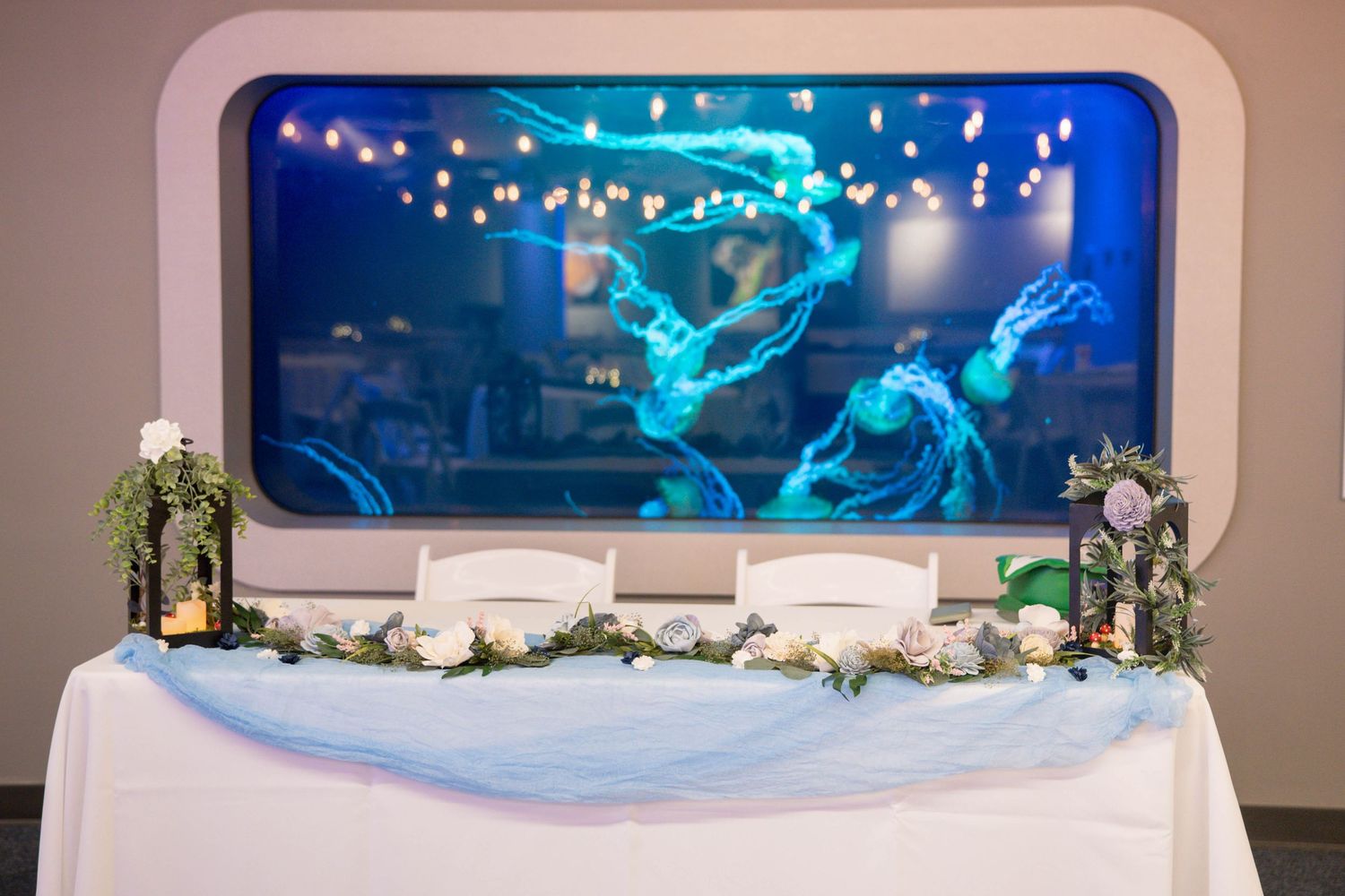 A wedding reception head table decorated with lights and featuring an aquarium display with blue lighting in the background.