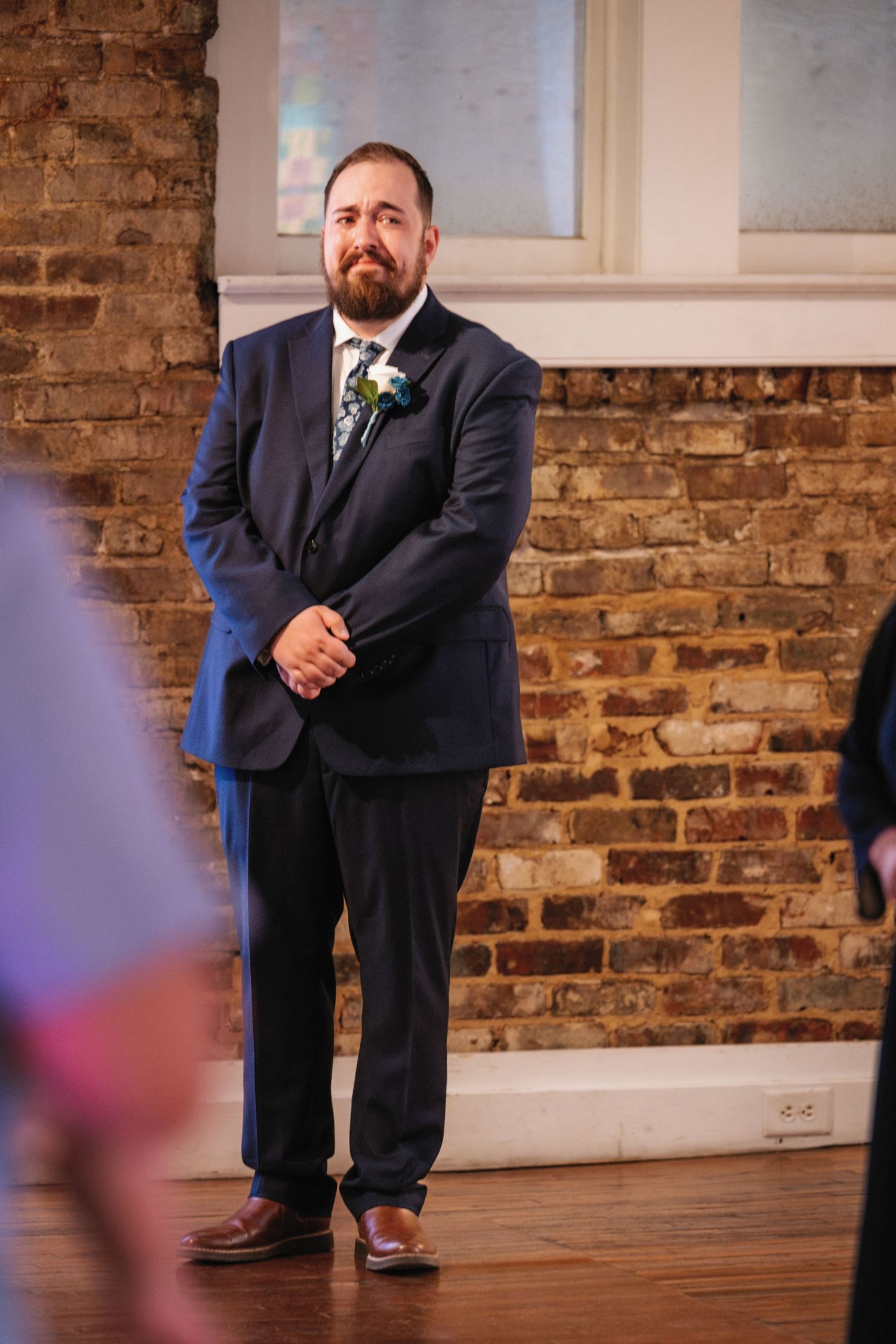 Someone in a navy suit stands against a rustic brick wall in what appears to be a wedding venue.