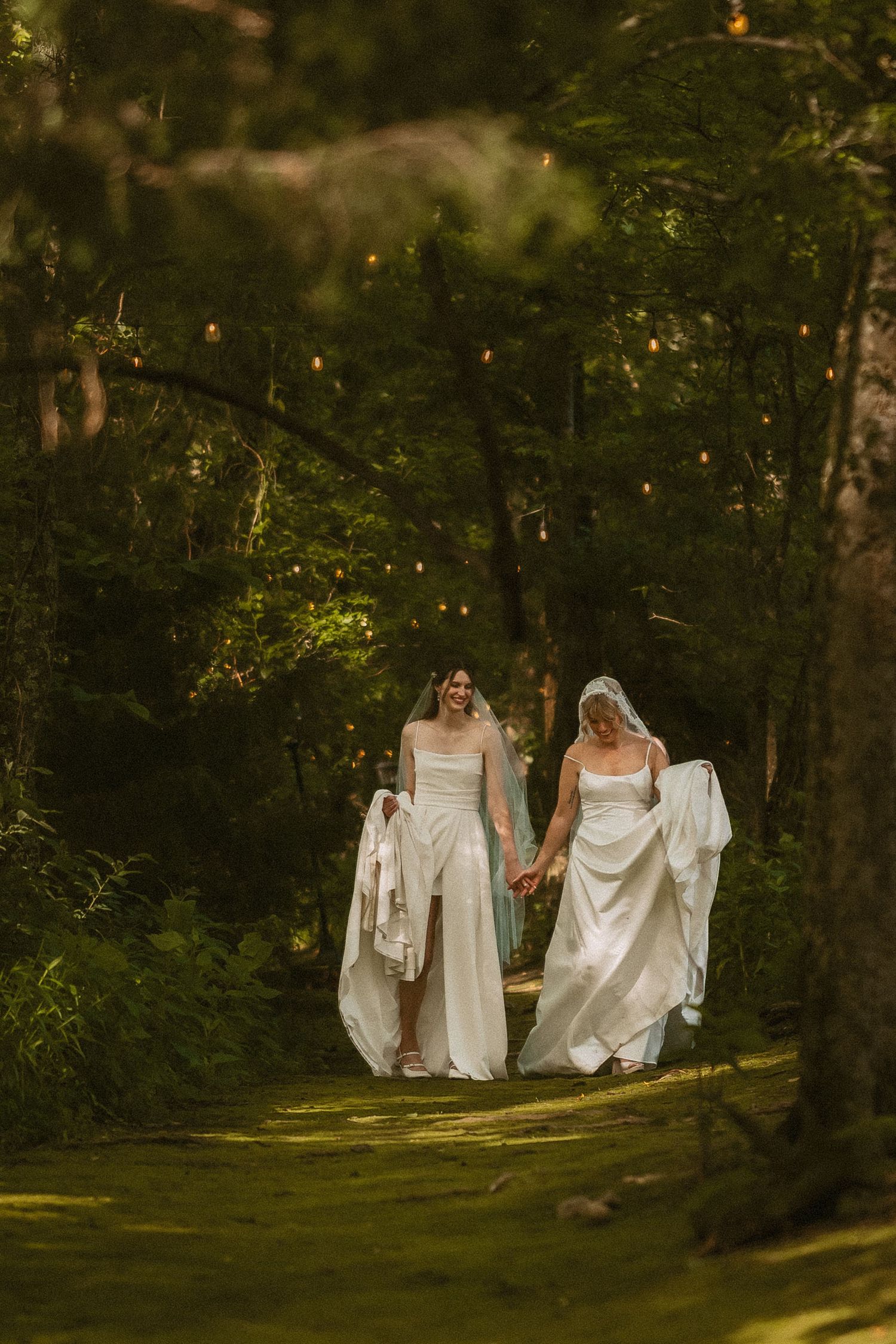 Two figures in flowing white dresses walk through a sunlit wooded path during golden hour.