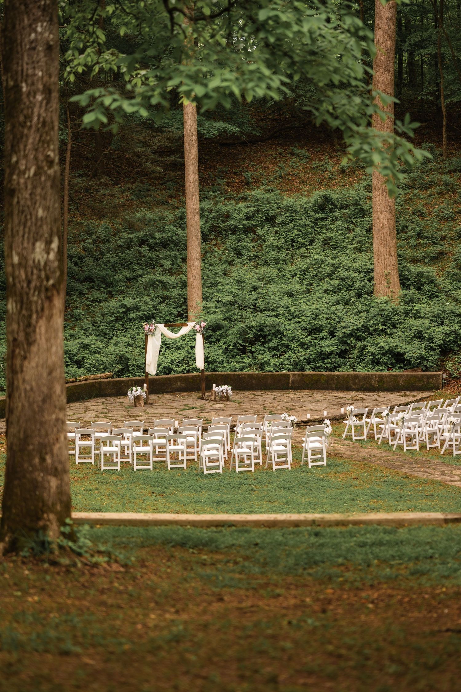 White chairs arranged for an outdoor forest wedding ceremony with a wooden arch decorated with fabric in a wooded setting.