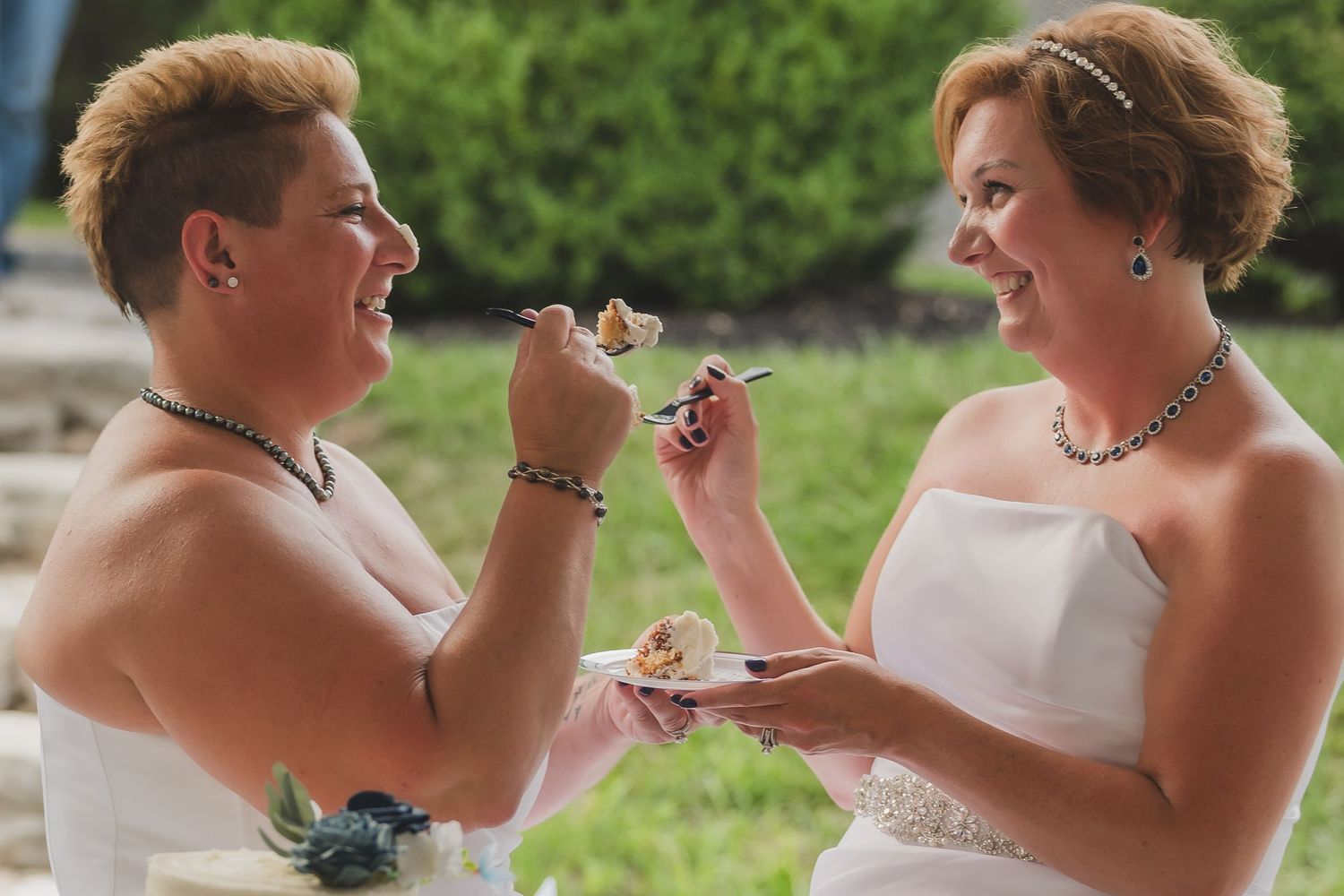 A couple in formal wedding attire playfully feed each other cake during their wedding reception celebration outdoors.