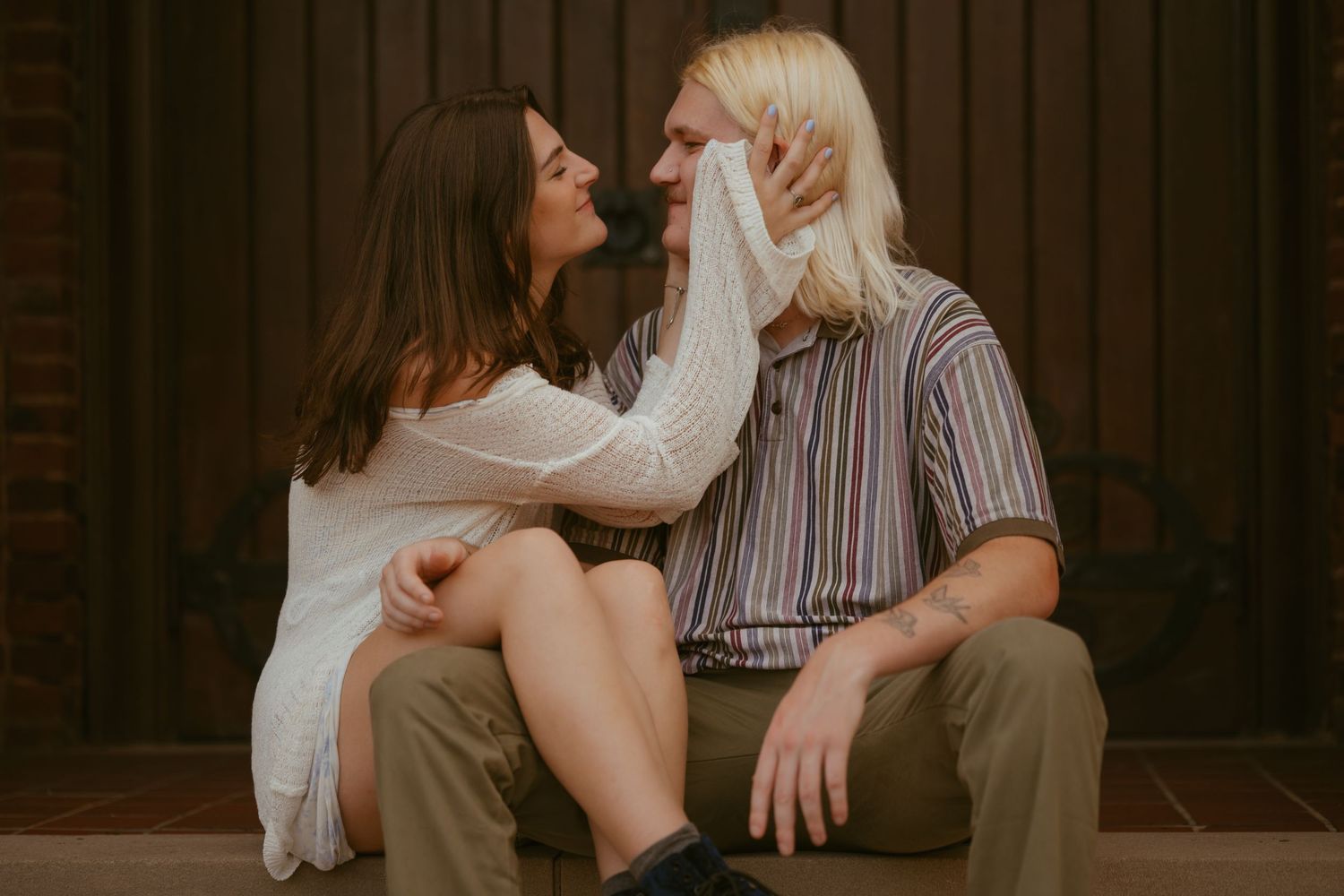 A couple in casual attire shares an intimate moment while sitting on wooden steps in a dimly lit setting.