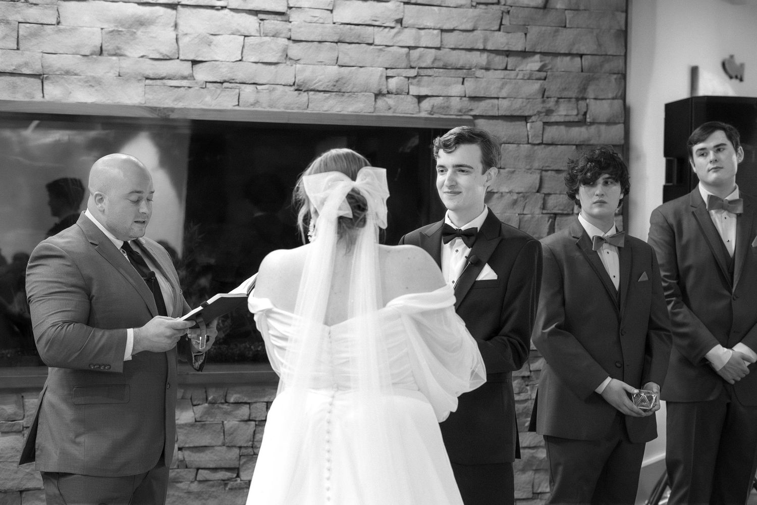 Black and white wedding ceremony photo showing bridal party gathered in front of stone fireplace.