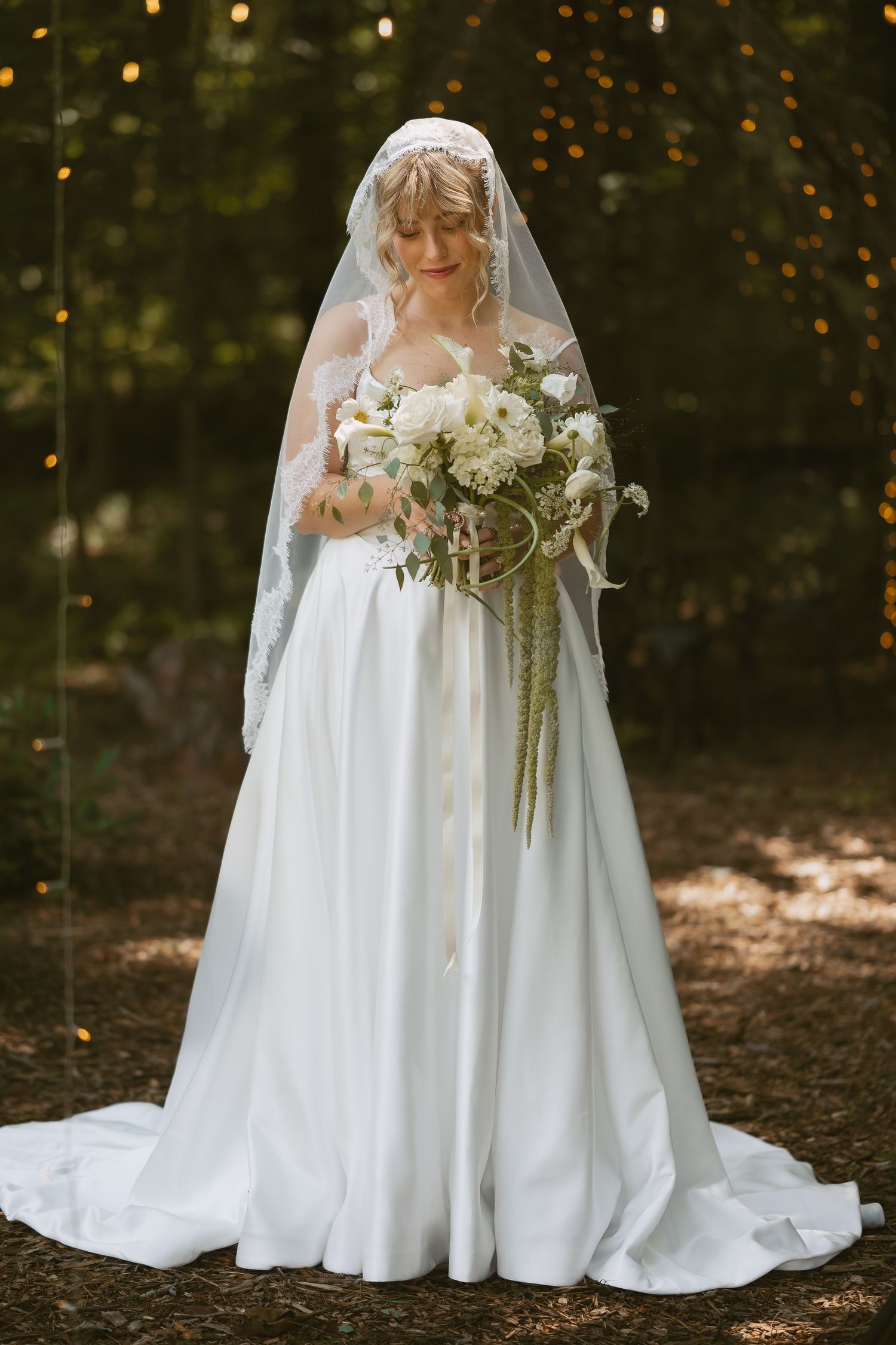 A bride in a flowing white gown holds a cascading bouquet while standing in a sun-dappled forest setting.