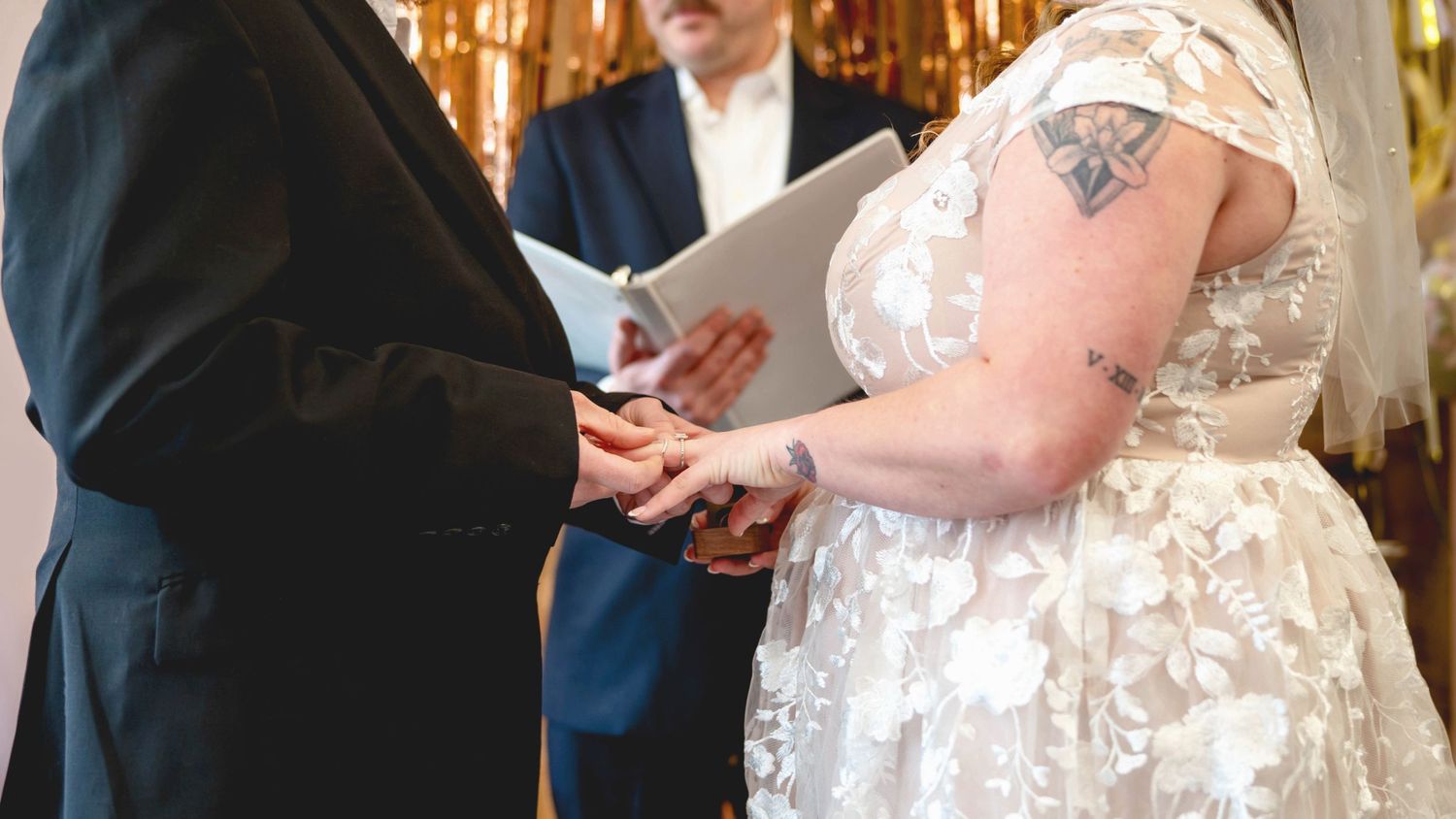 A couple exchanges rings during a wedding ceremony as an officiant presides, with the bride wearing a white lace gown.