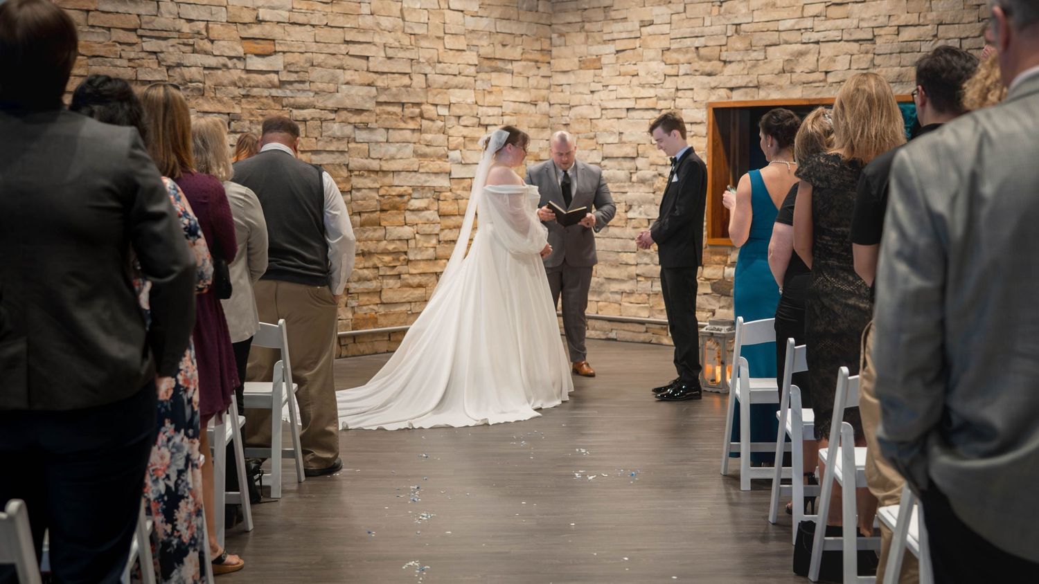 Intimate wedding ceremony taking place in rustic stone-walled venue with guests seated in white chairs.
