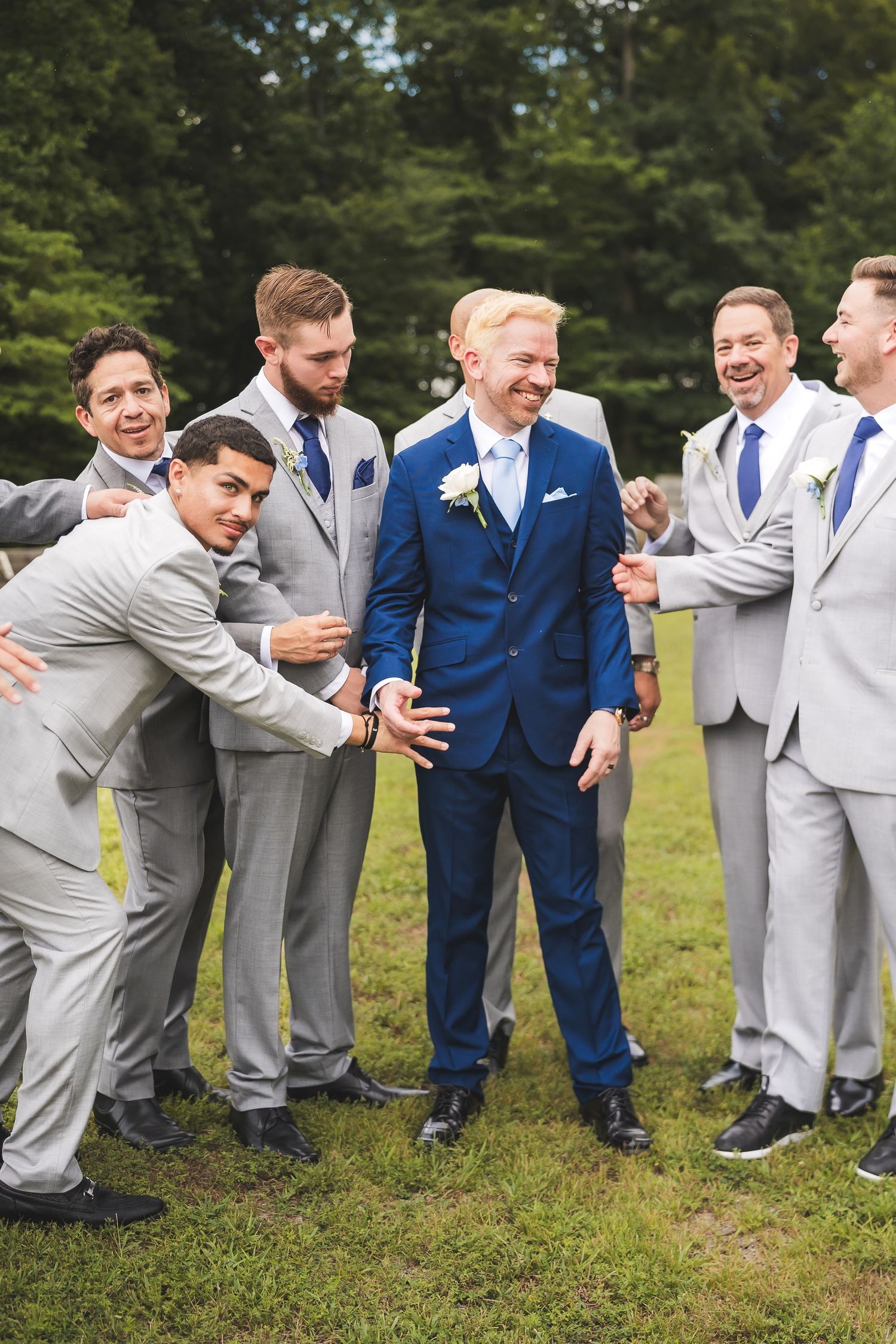 Groomsmen in grey and navy suits pose for a fun group photo while leaning on each other outdoors on grass.