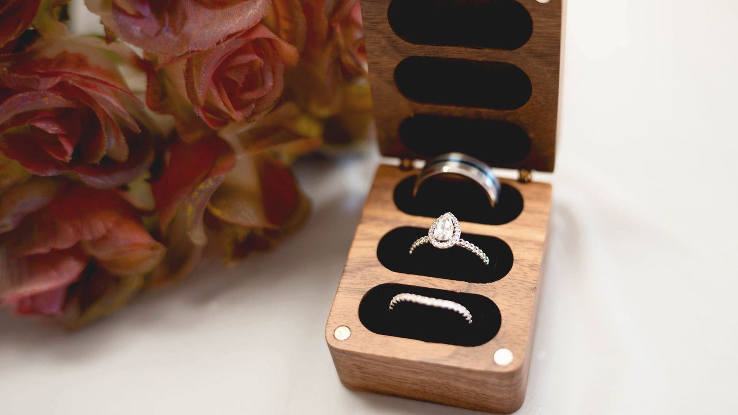 Rose gold ring boxes display elegant jewelry settings against black velvet cushions with pink roses in background.