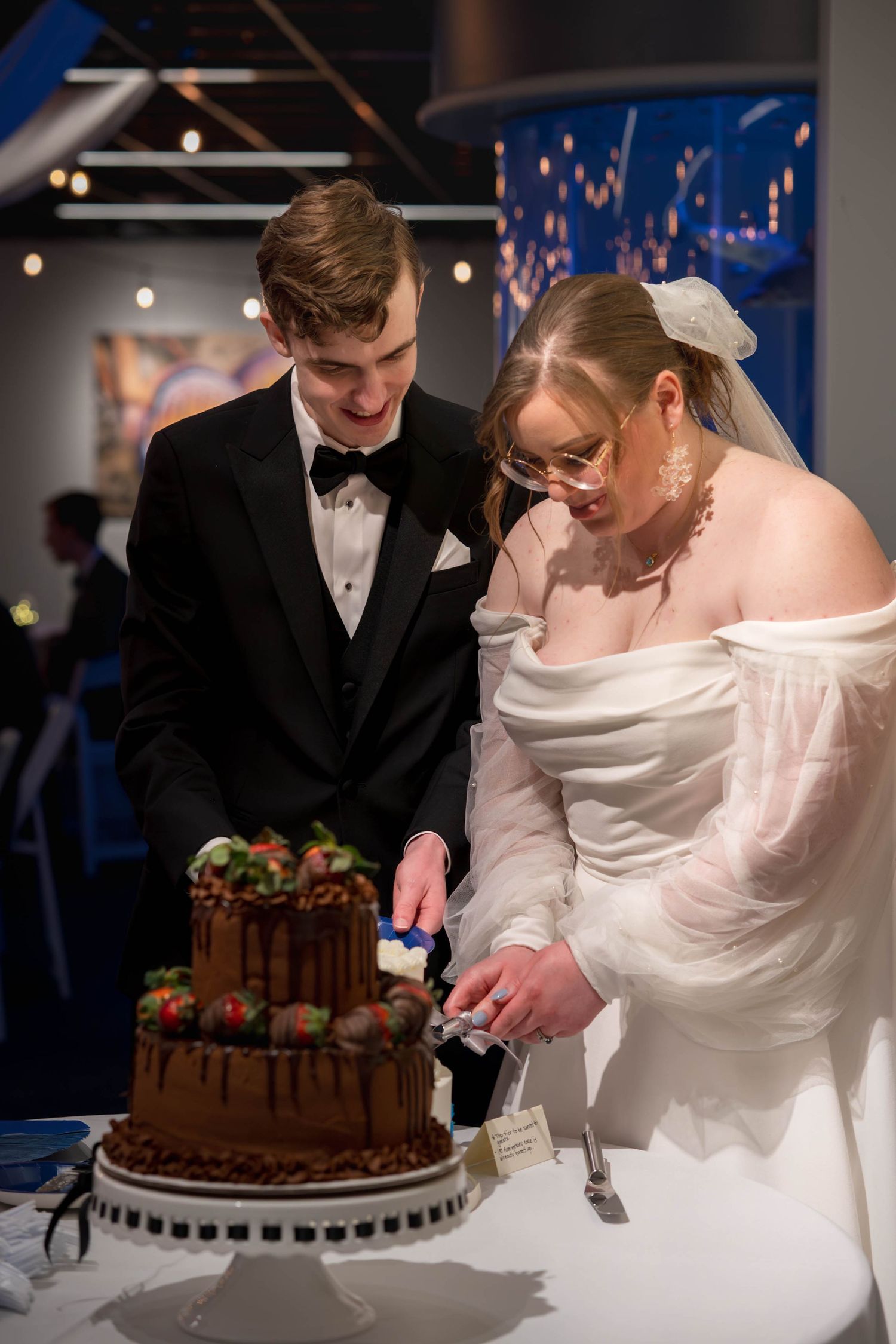 A newlywed couple cuts into their elegant three-tier chocolate wedding cake adorned with fresh strawberries.