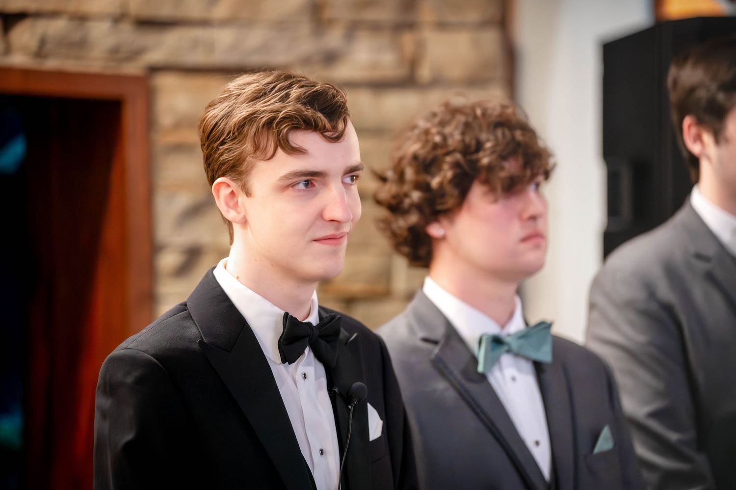 Two men in formal black tuxedos and bow ties at a wedding event, one wearing a mint colored tie.