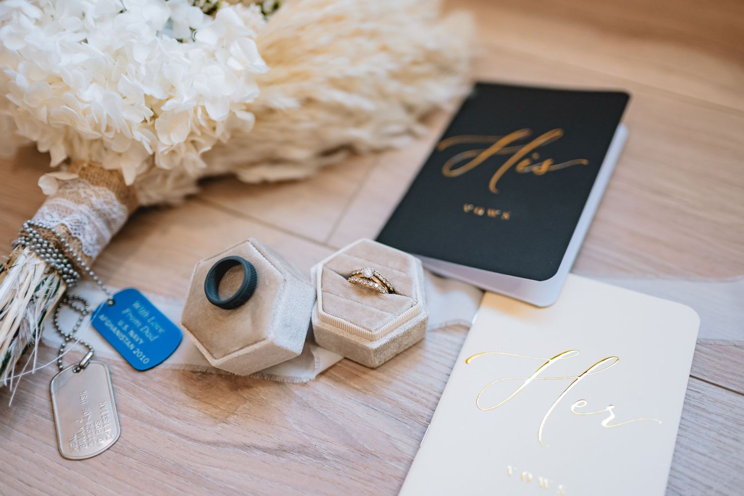 Wedding details including rings in hexagonal boxes, vow books, and white floral bouquet arranged on wooden surface.