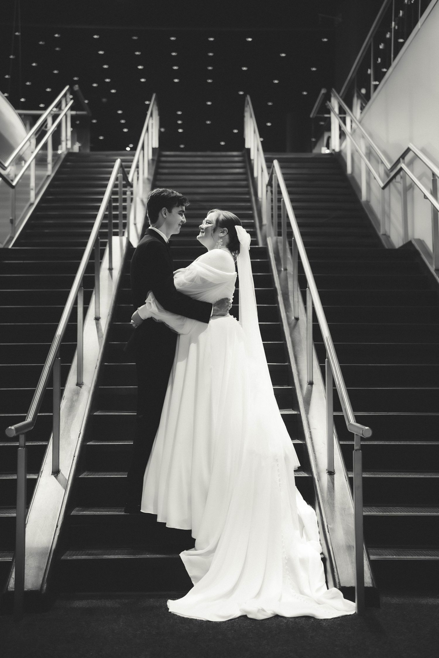Elegant bride and groom share an intimate moment on modern stairs with dramatic lighting.