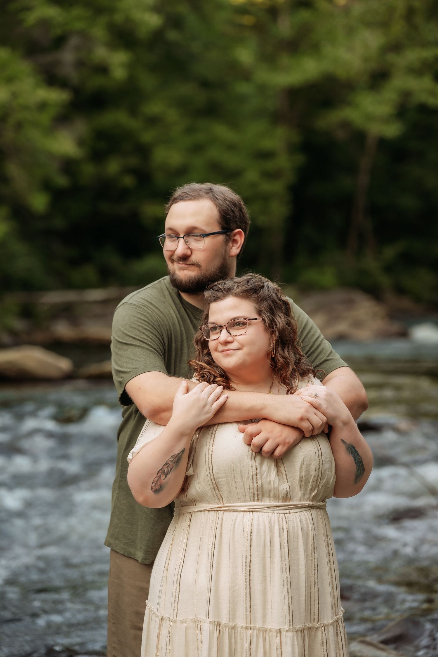 A couple embraces by a flowing river surrounded by lush greenery during an outdoor photoshoot.