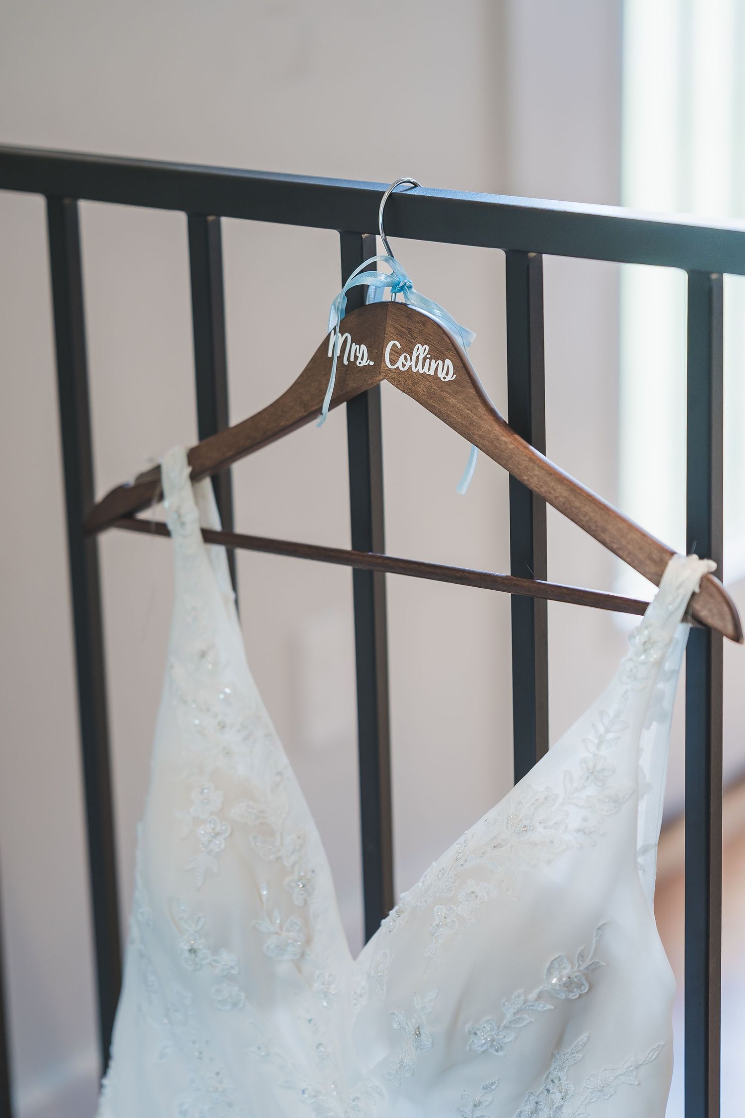 A white lace wedding dress hangs on a wooden hanger labeled 'Mrs. Collins' with a blue ribbon accent.