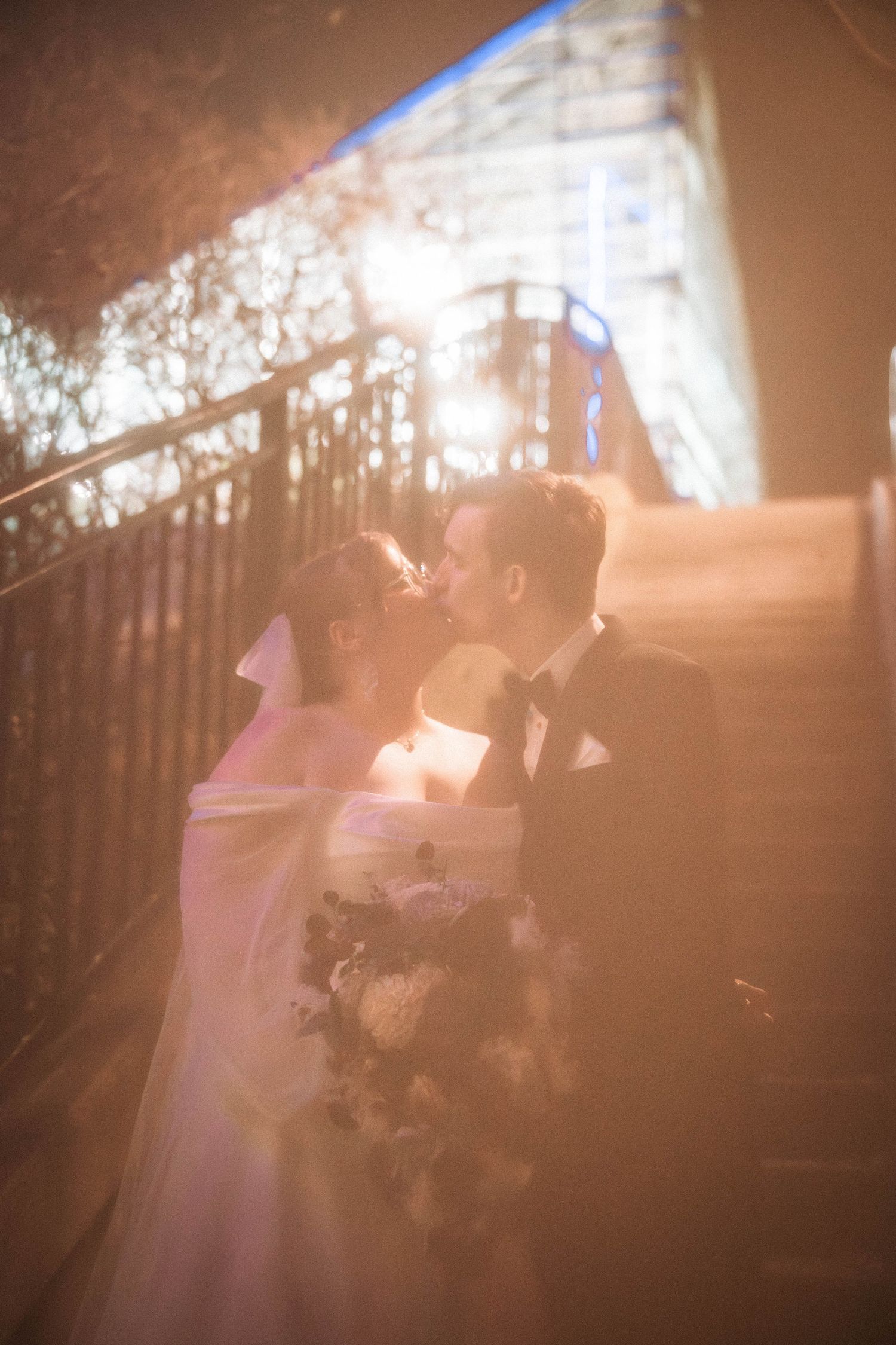 A romantic wedding couple shares an intimate kiss on a staircase with soft lighting and a floral bouquet.