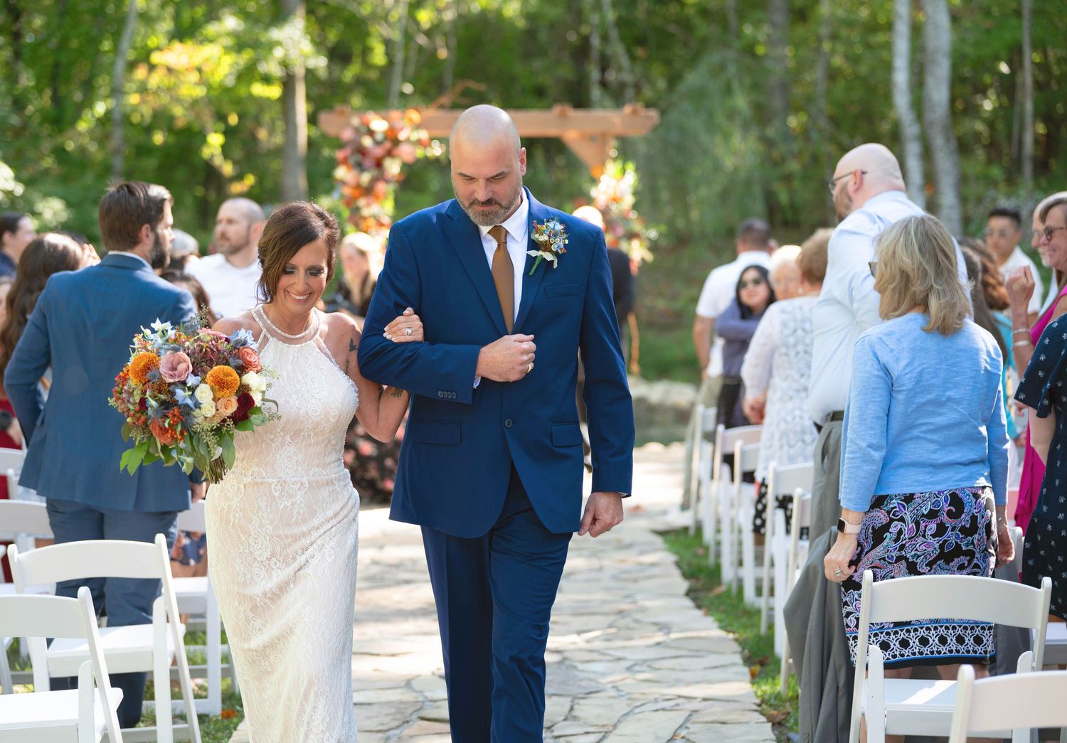 Outdoor wedding ceremony processional with guests seated in white chairs along a stone pathway.