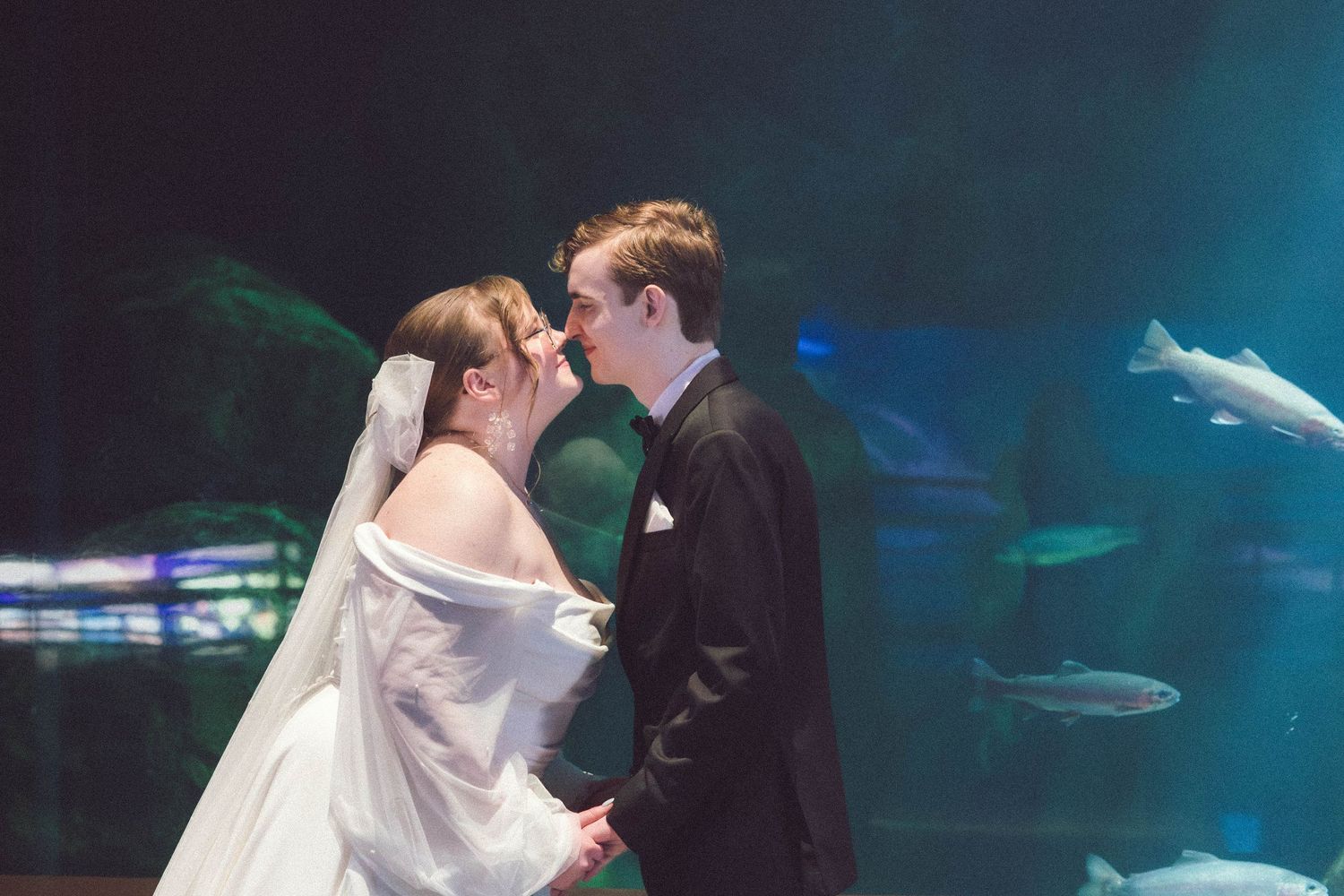 A bride and groom share a romantic kiss in front of a glowing aquarium tank with ethereal blue lighting.