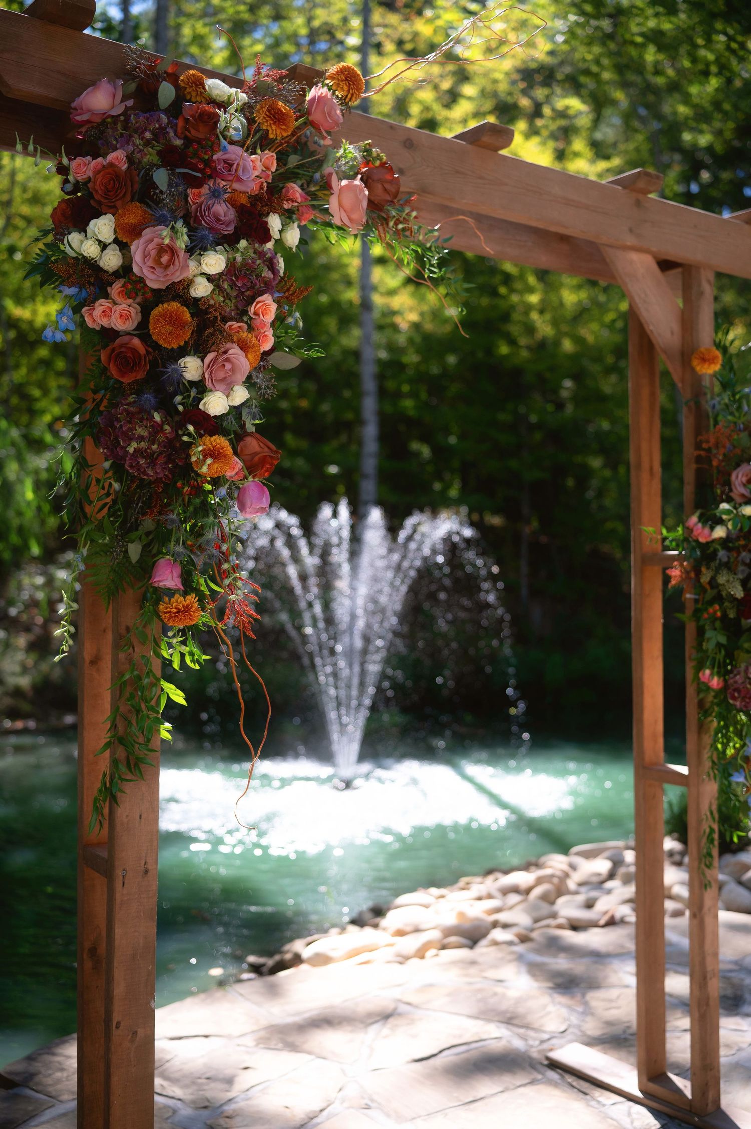 Wooden wedding arch decorated with pink and orange flowers stands before a serene fountain in a garden setting.