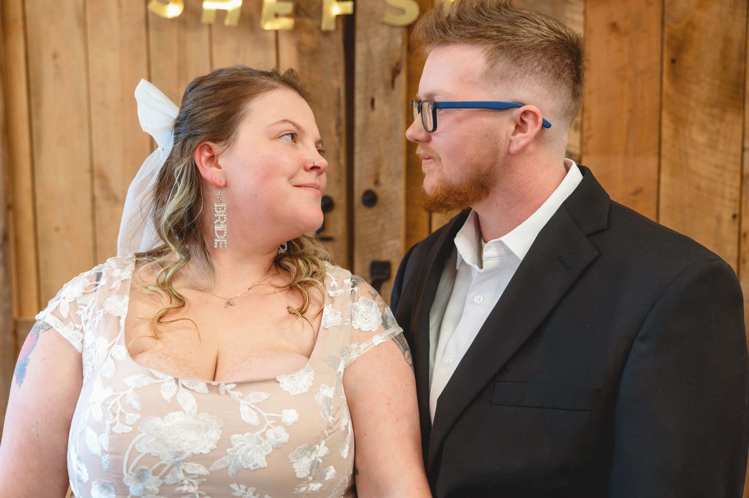 Two people share a loving look at their wedding, with the bride in a lace gown and groom in a black suit inside a barn setting.