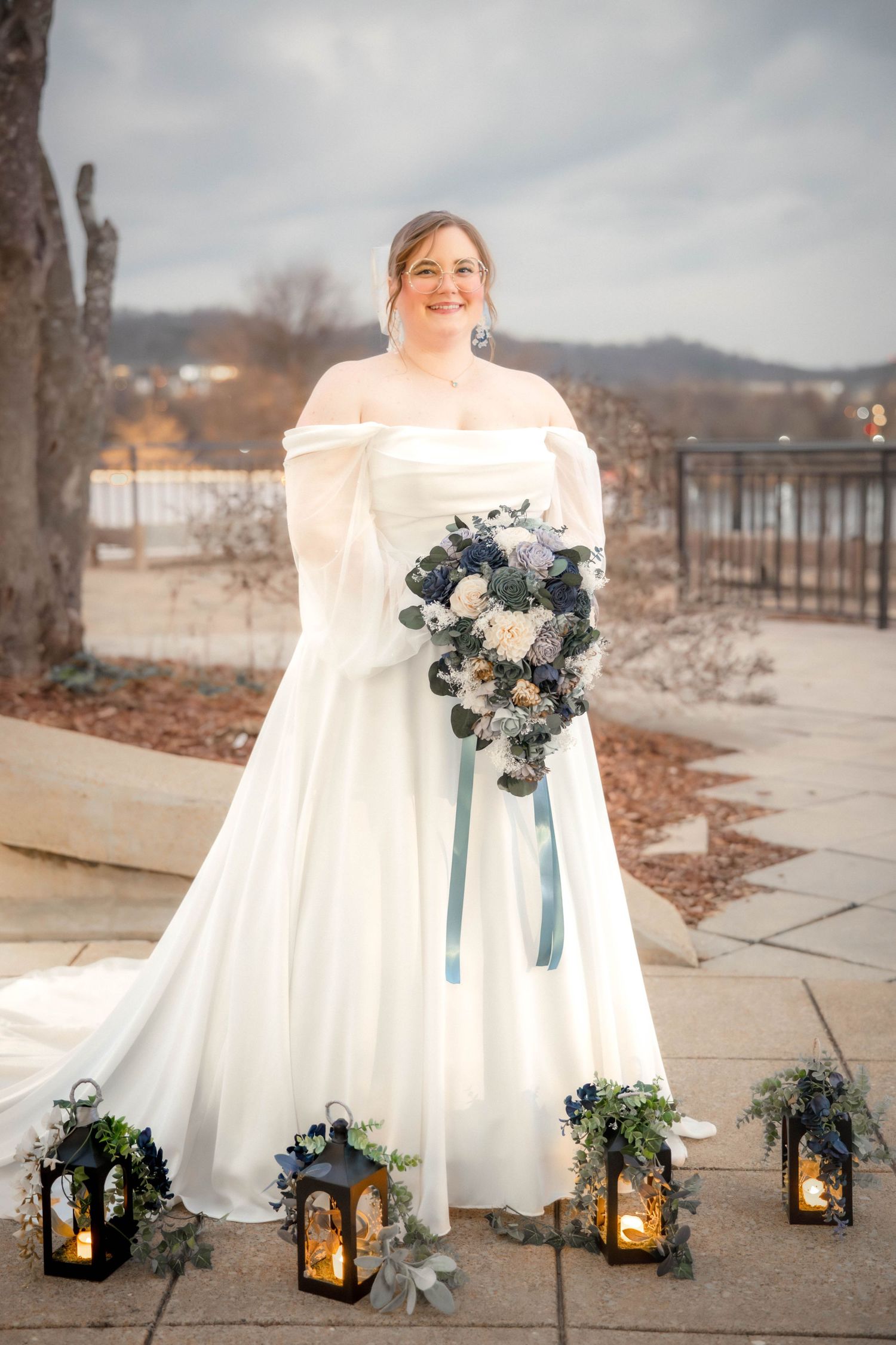 A winter wedding scene with an elegant off-shoulder white gown, lantern decor and a blue and white bouquet on a lakeside path.