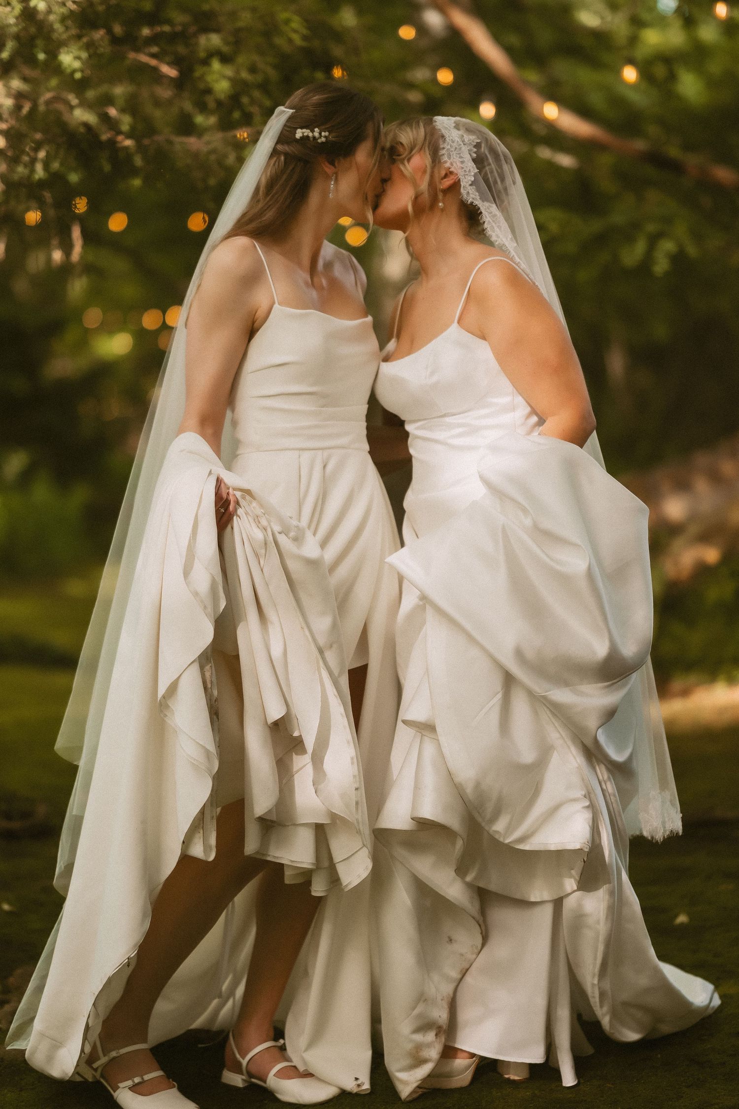Two flowing white wedding dresses captured in elegant outdoor garden setting with bokeh lights.