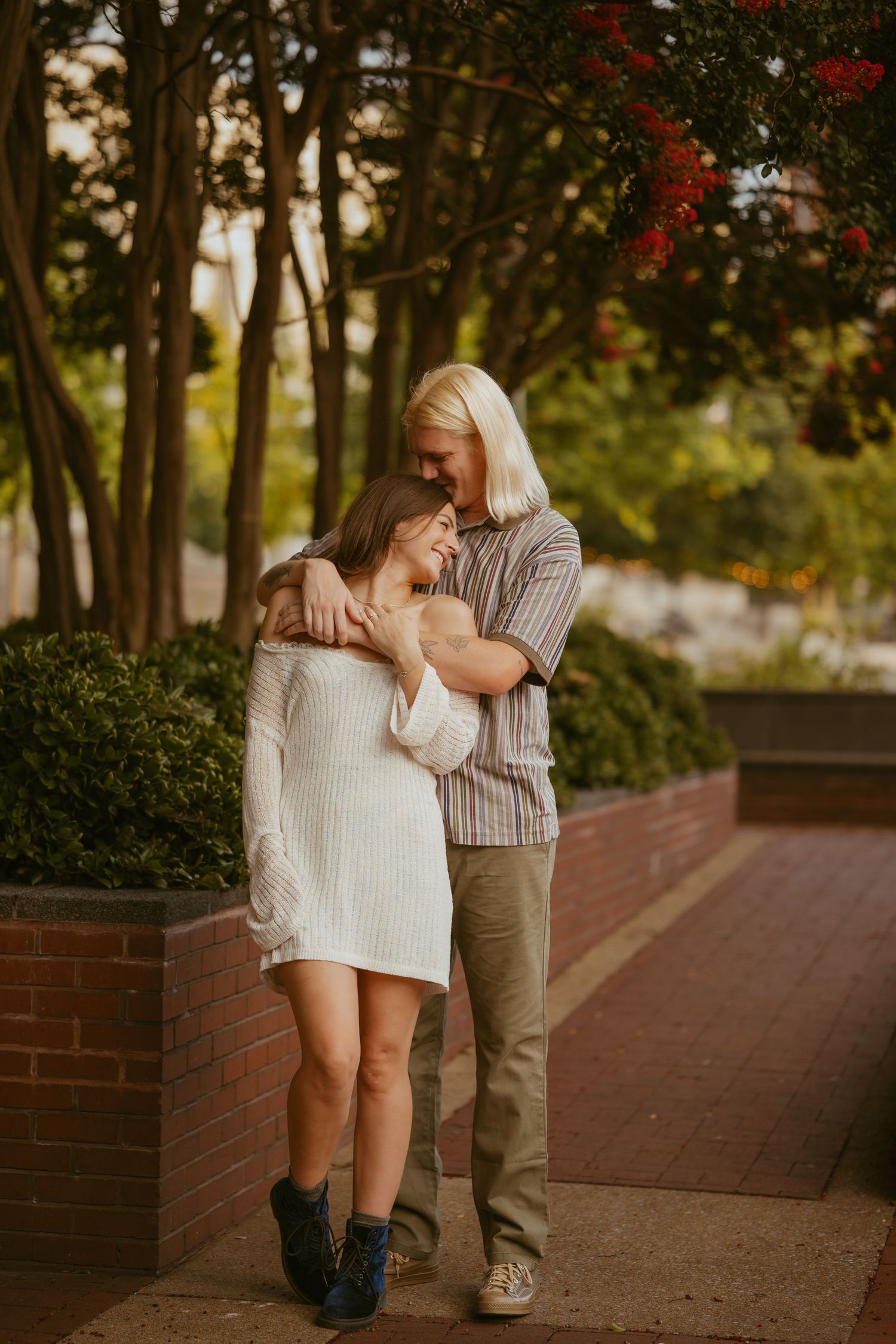 A couple shares an intimate moment outdoors, embracing beside a brick planter in warm sunset lighting.
