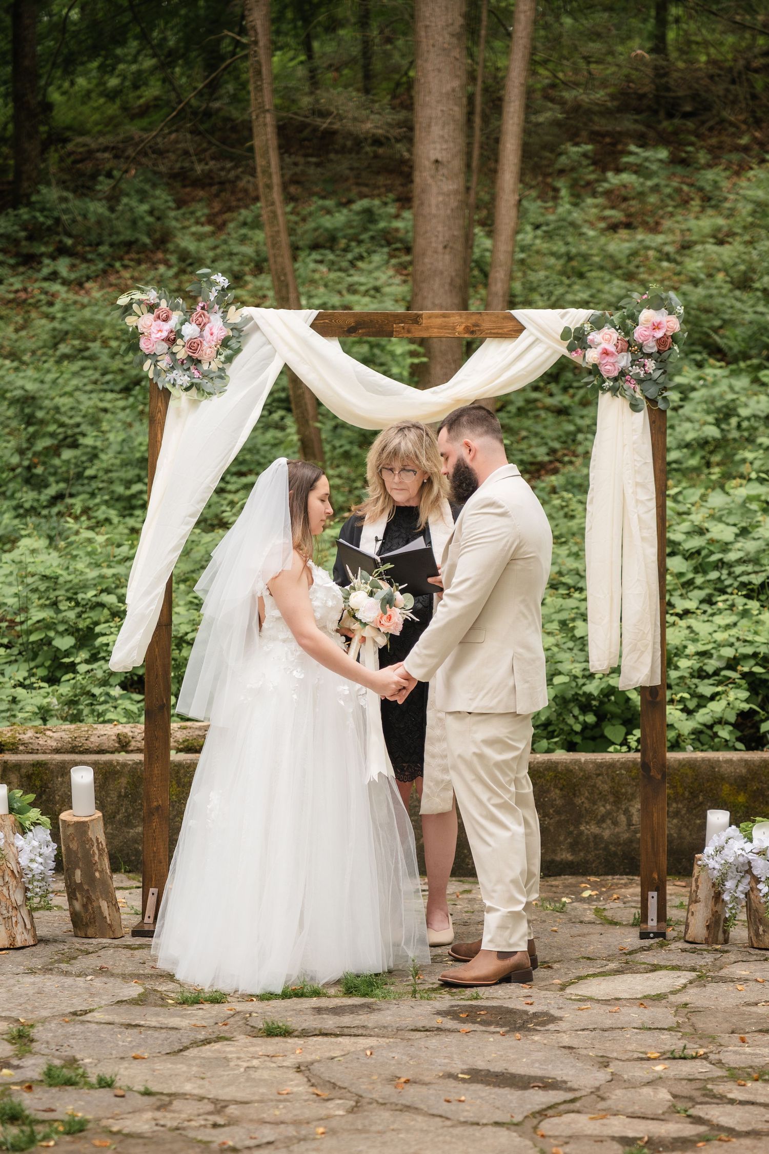 A romantic outdoor wedding ceremony takes place under a white draped arch in a wooded forest setting.