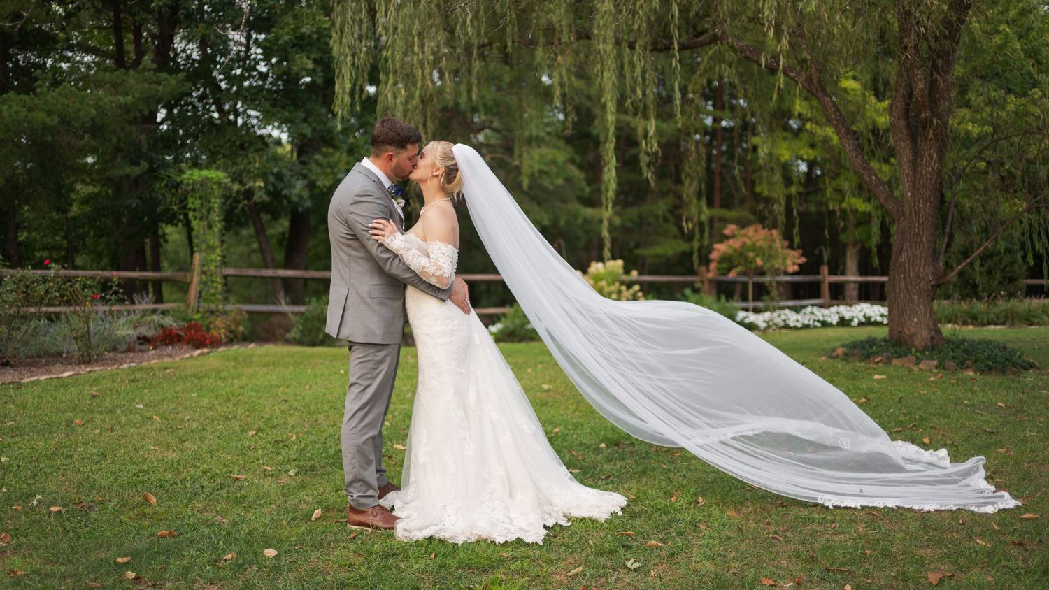 A long white wedding veil billows dramatically in the wind across a lush green garden setting.