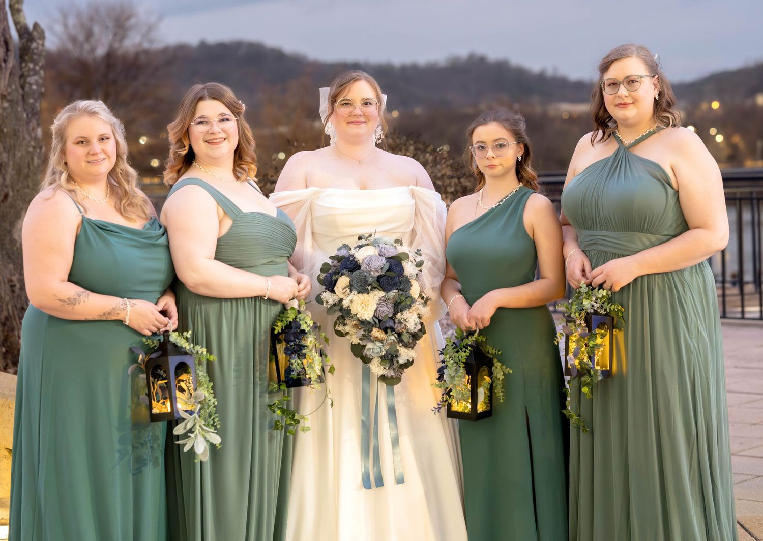 Bride in white gown stands with four bridesmaids wearing matching sage green dresses holding lantern bouquets.