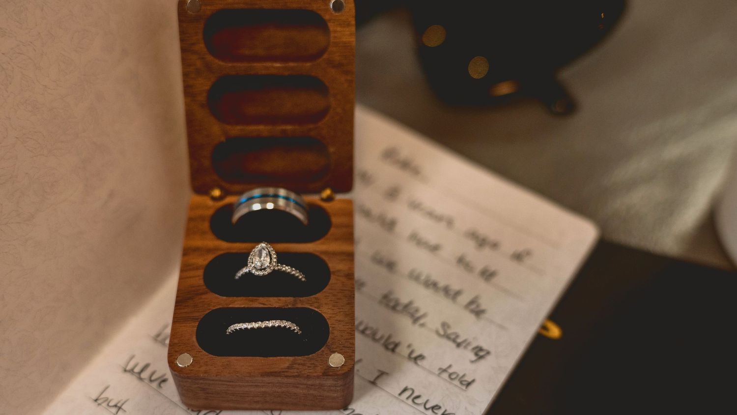 A vintage wooden ring box sits open on handwritten paper, featuring a traditional diamond engagement ring.