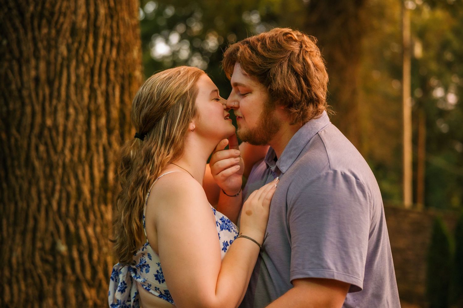 A romantic couple share an intimate embrace while standing near a tree trunk during golden hour.