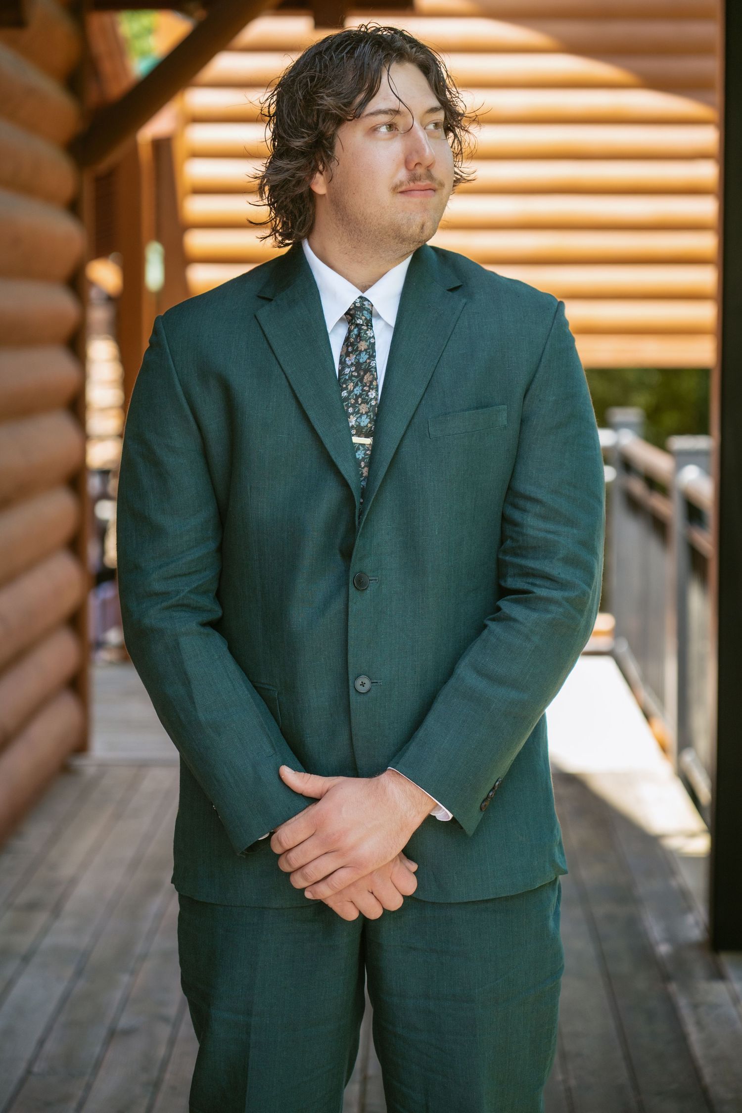 A person in a dark green suit stands in front of wooden panels with natural lighting.