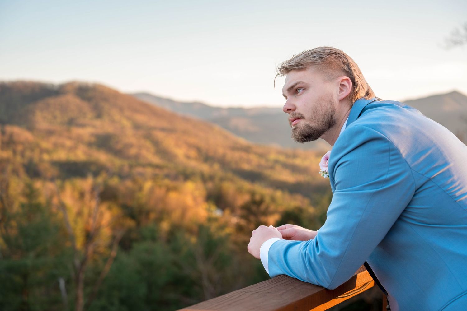 A person wearing a blue jacket stands on a mountain trail overlooking scenic autumn foliage during golden hour.