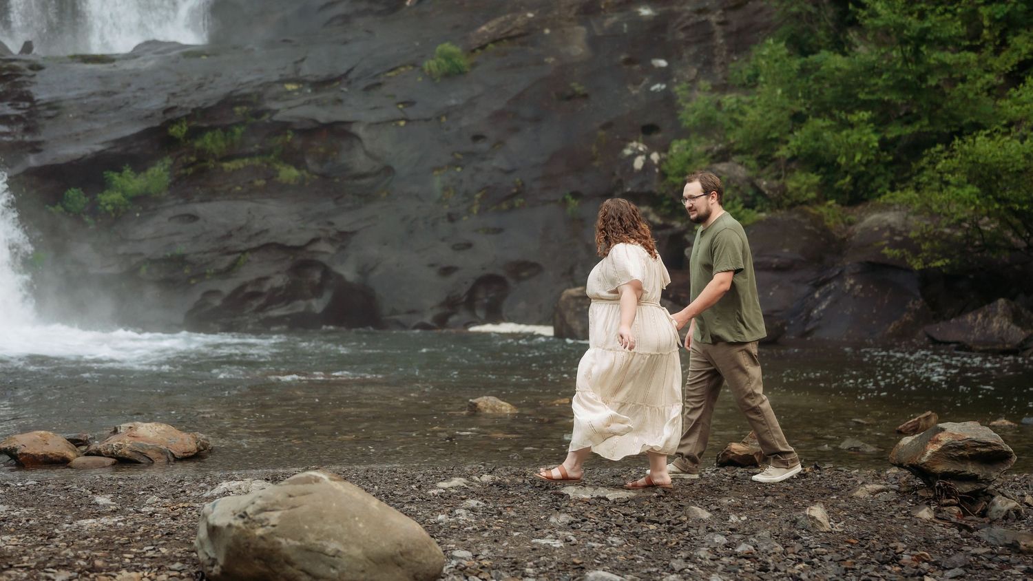 A couple holds hands while walking near a scenic waterfall on rocky terrain surrounded by lush greenery.
