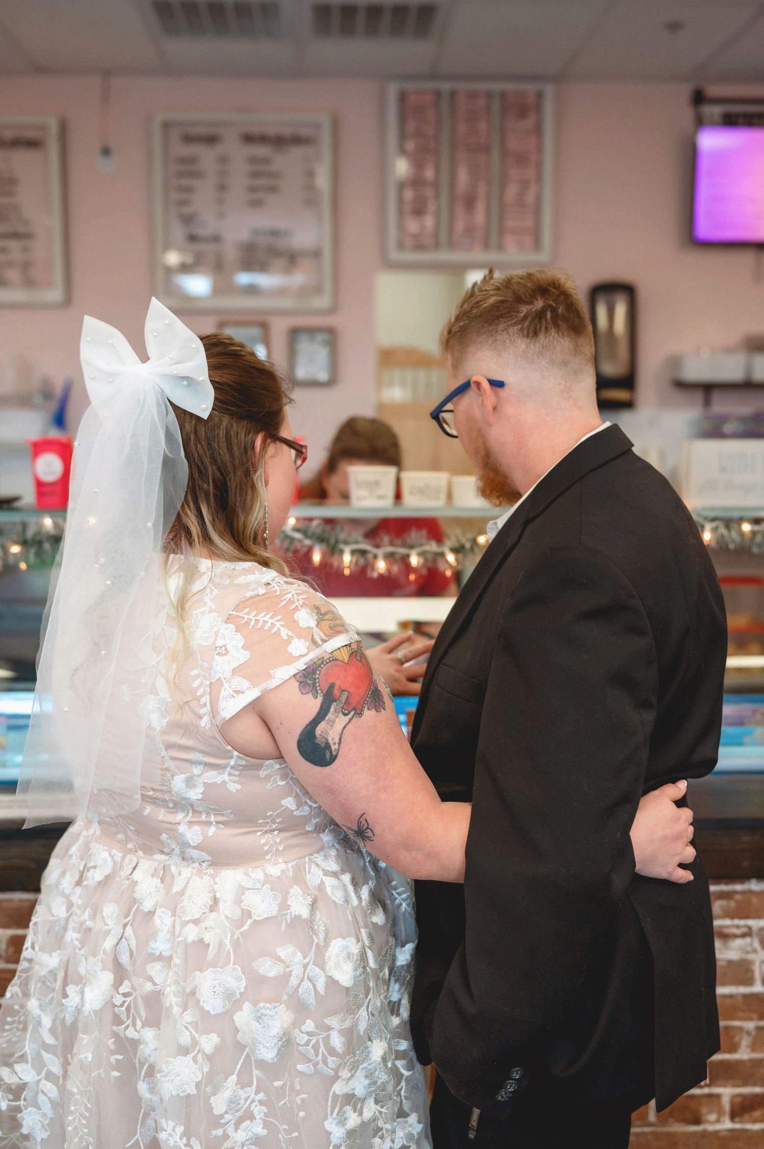 A couple in wedding attire stand inside an ice cream shop near a glass display case.