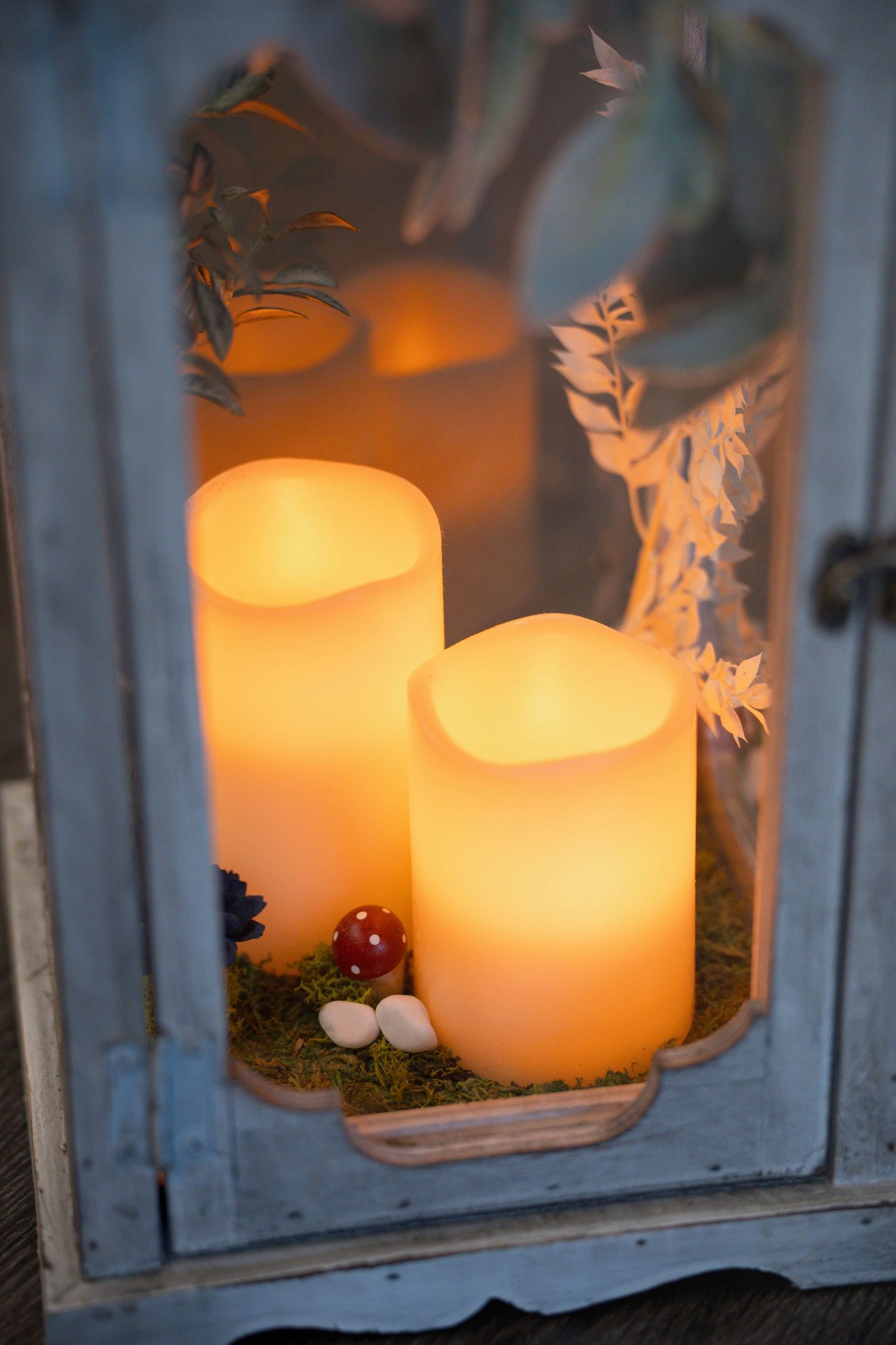 Two glowing LED pillar candles in a rustic lantern display with decorative moss and small mushroom accents.